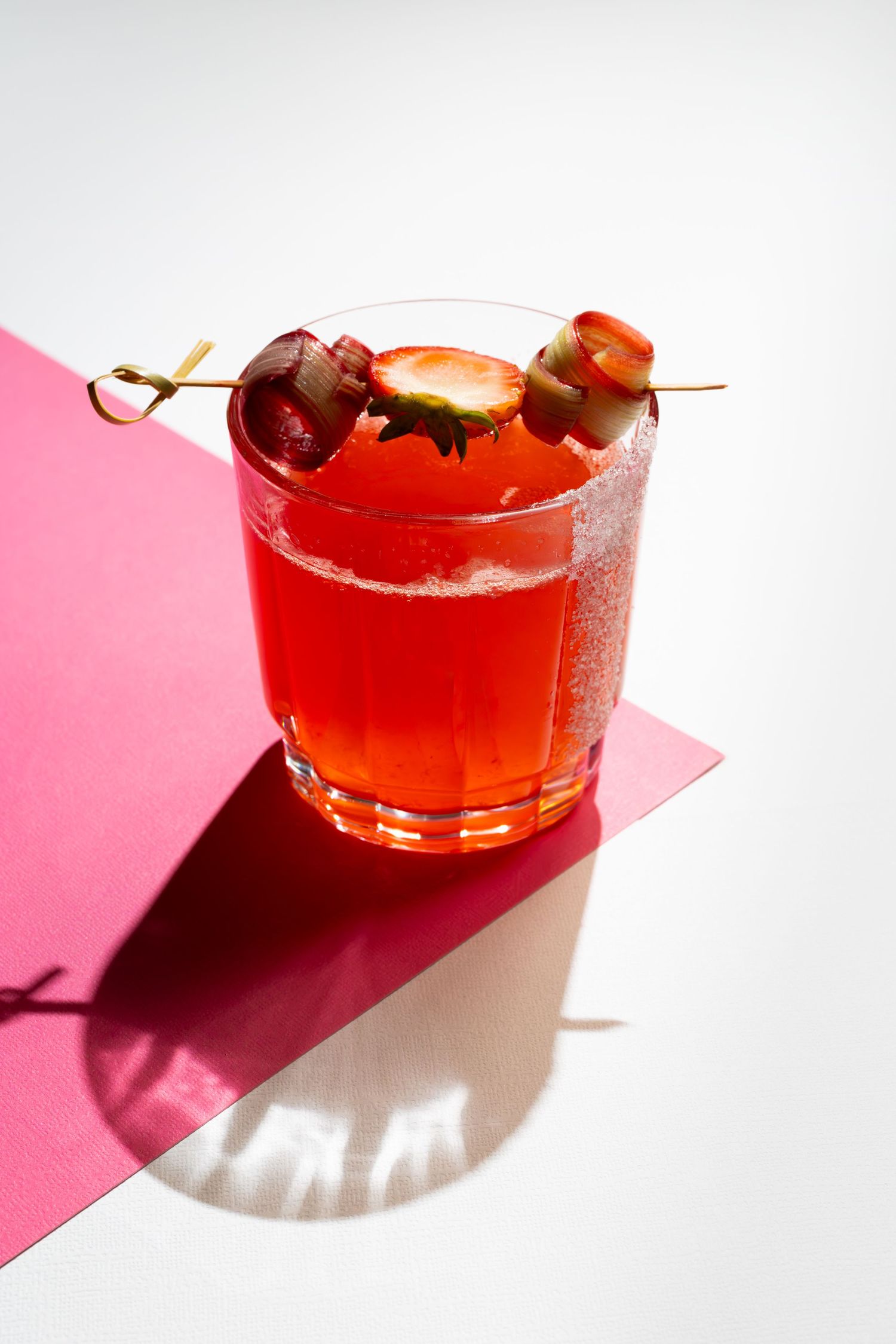 Non-alcoholic sugared rimmed strawberry rhubarb mocktail with fresh fruit garnish on white and pink backdrop. B. Steele Images