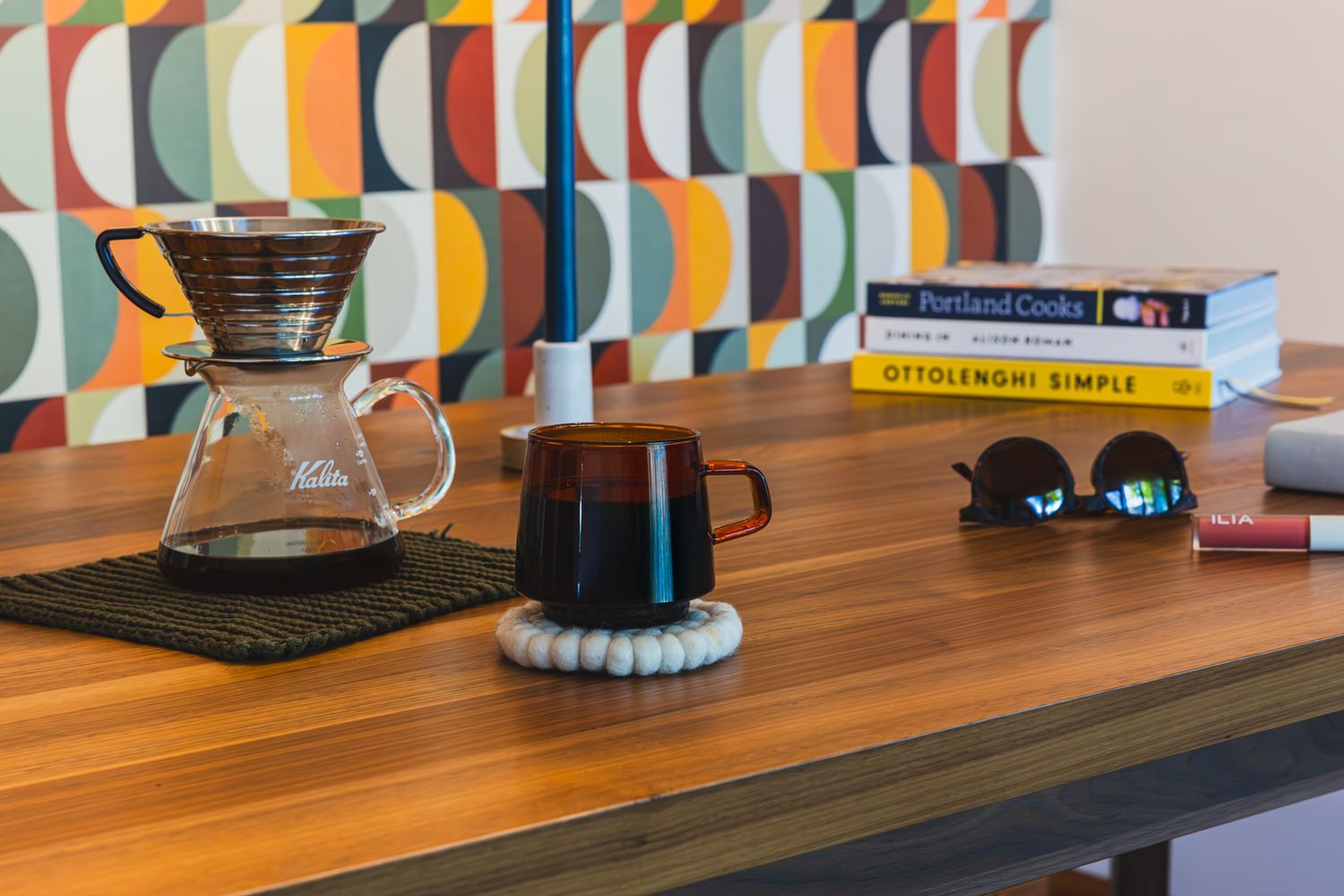 A Kalita pour-over coffee maker and mug on a wooden desk beside stacked books and sunglasses with a colorful geometric wall.