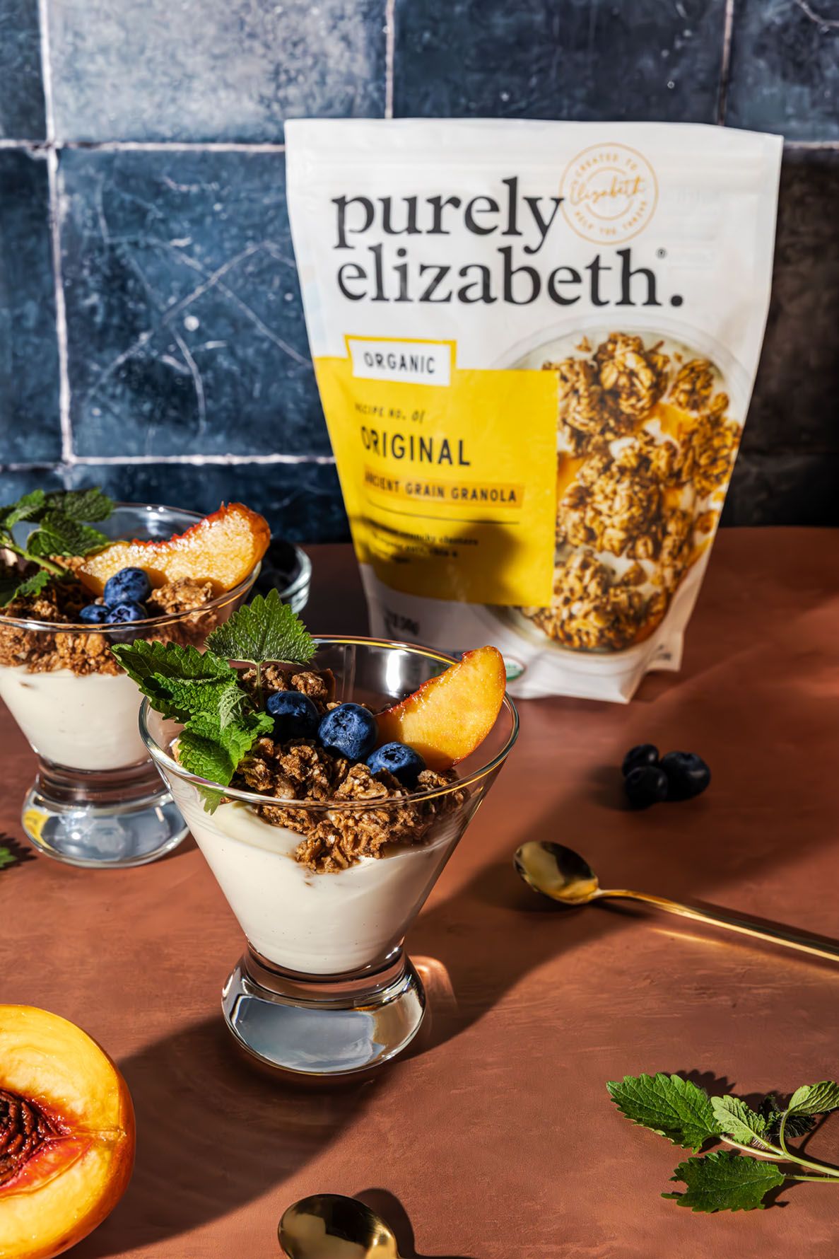 Yogurt parfait cups with Purely Elizabeth granola and fresh fruit arranged in a row on a dark wooden surface with mint garnish.