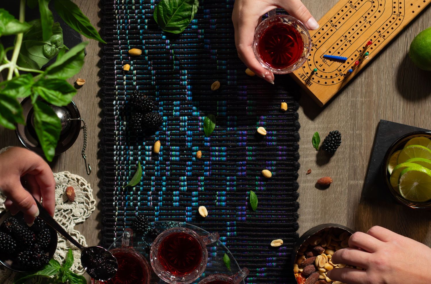 Top-down view of hands around placemats with crystal glasses, snacks and greenery on a dark wooden table.