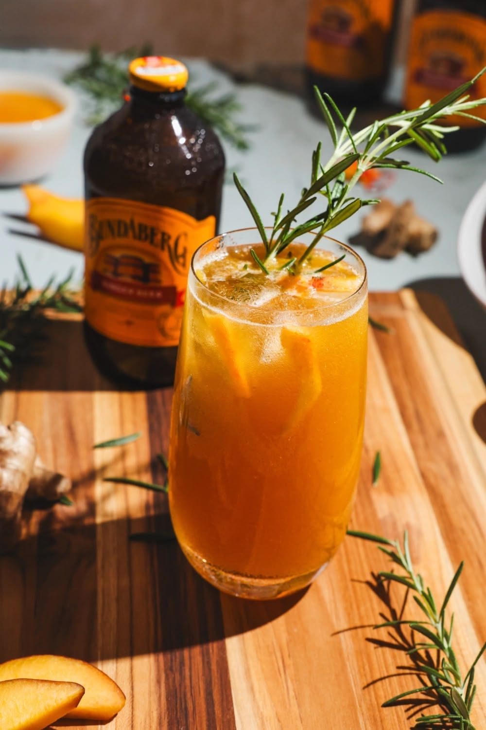 Orange Bundaberg mocktail garnished with fresh rosemary sprig served in a coupe glass on wooden surface with bottle in background.
