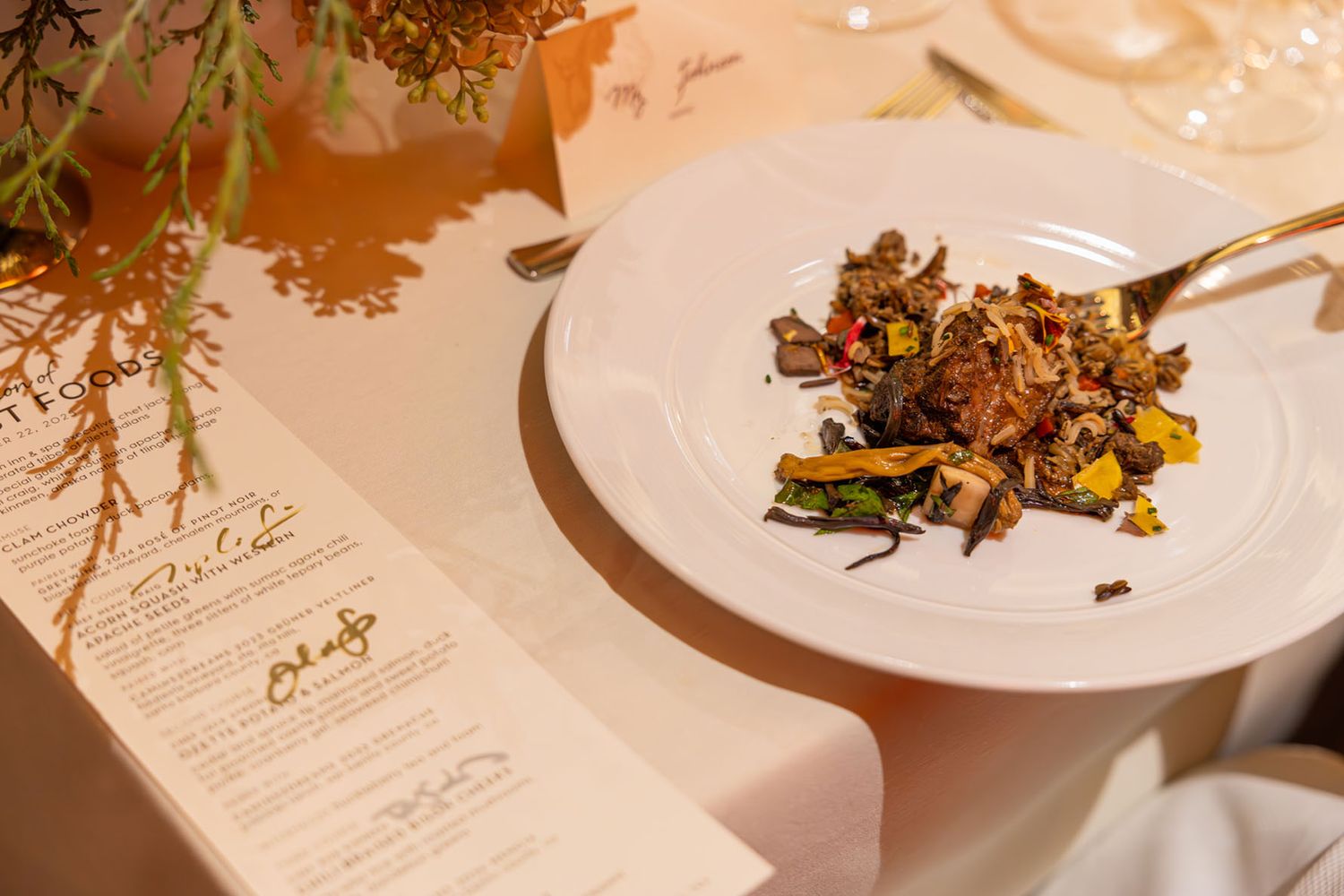 Elegant table setting with white plates, floral arrangements, and menu cards in warm lighting at an upscale event.