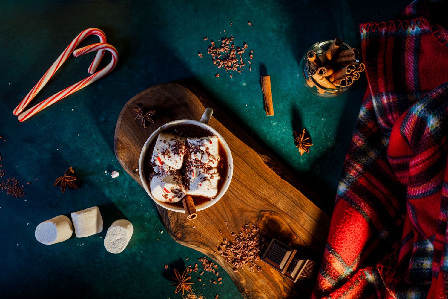 Hot chocolate topped with marshmallows and cinnamon sticks served in a glass mug with cozy plaid blanket and candy canes