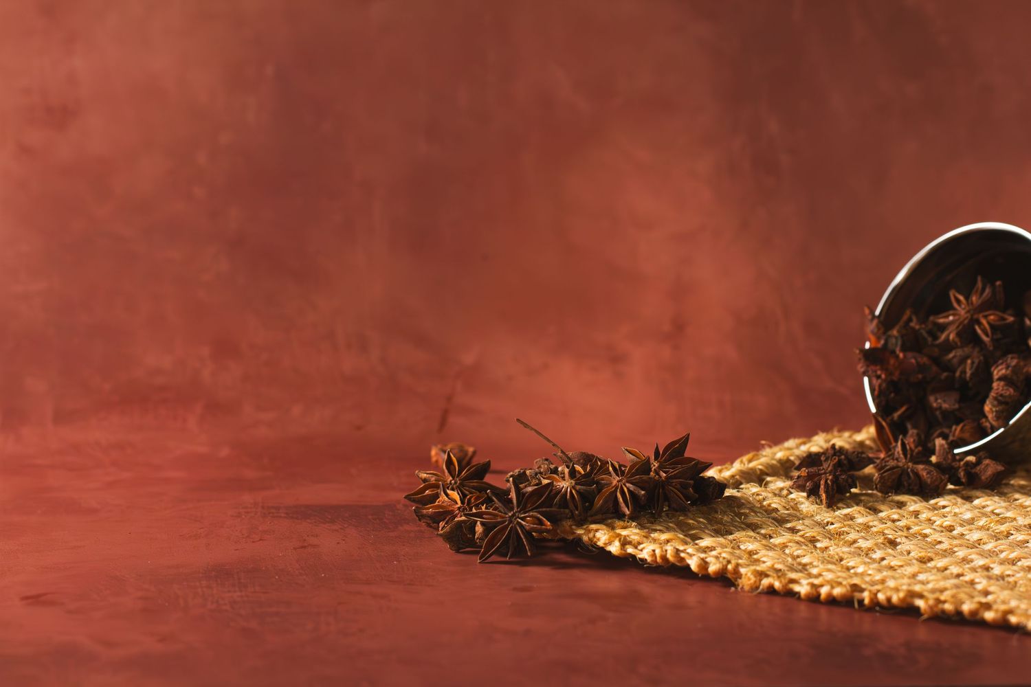 Scattered star anise spices spilled from a container onto a burlap fabric against a dark red background.
