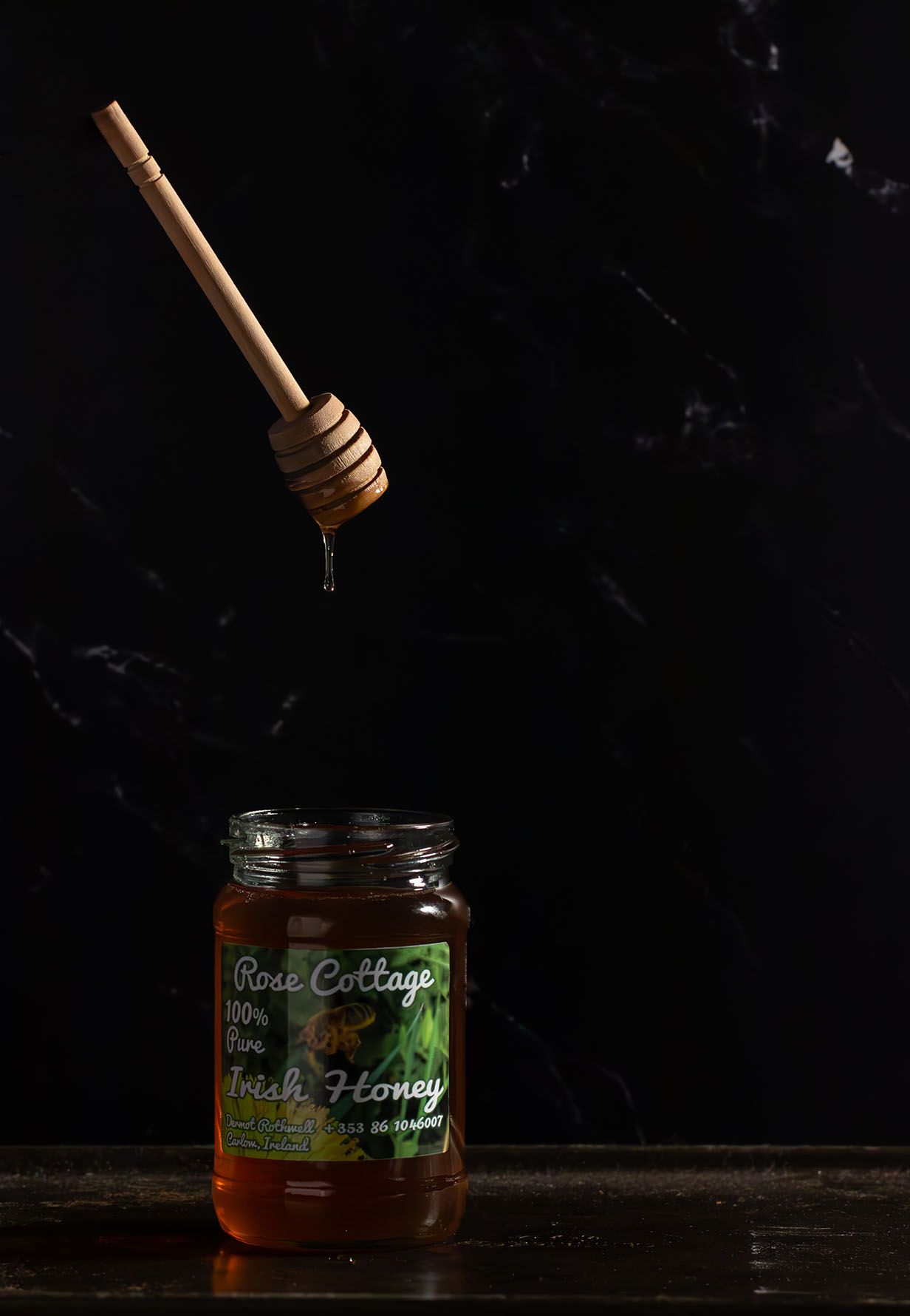 A wooden honey dipper drips golden honey from a glass jar against a dark background in moody lighting.