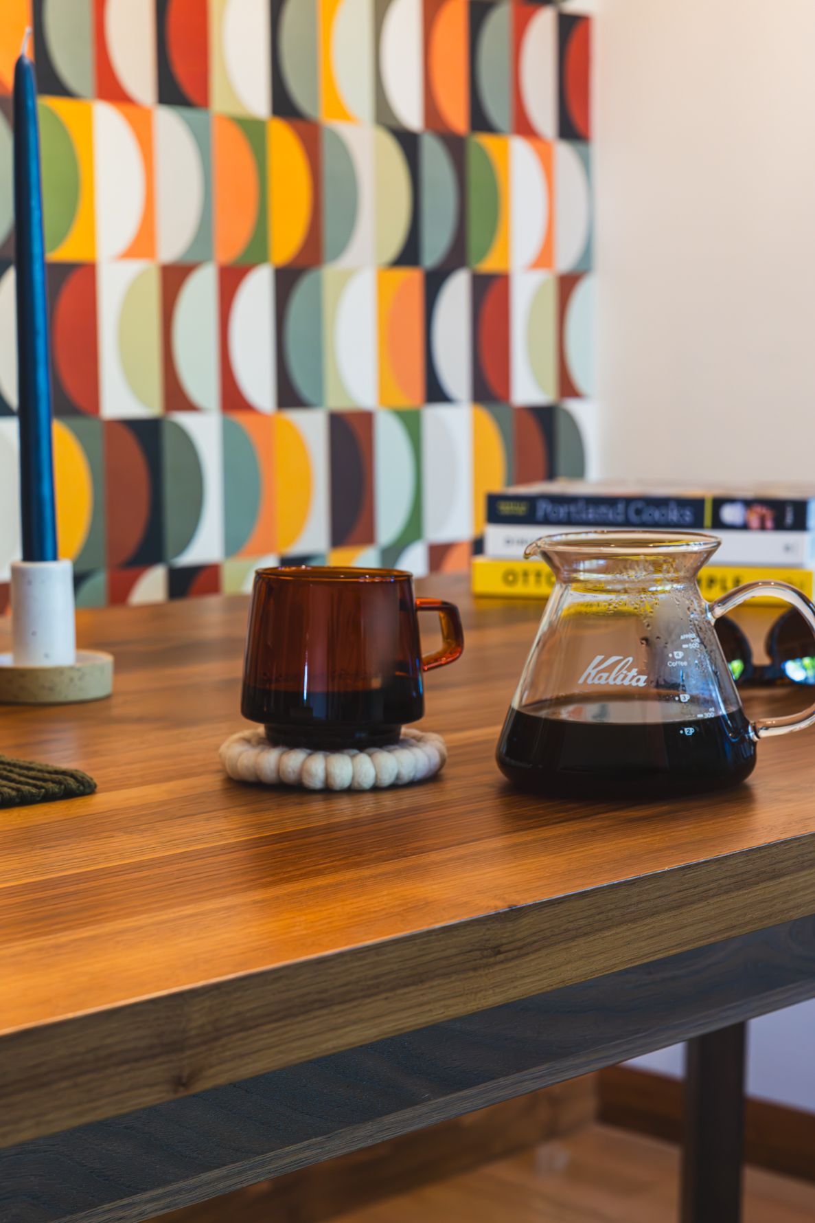 A Kalita Wave pour-over coffee maker and amber glass mug on a wooden table with colorful geometric art in the background.