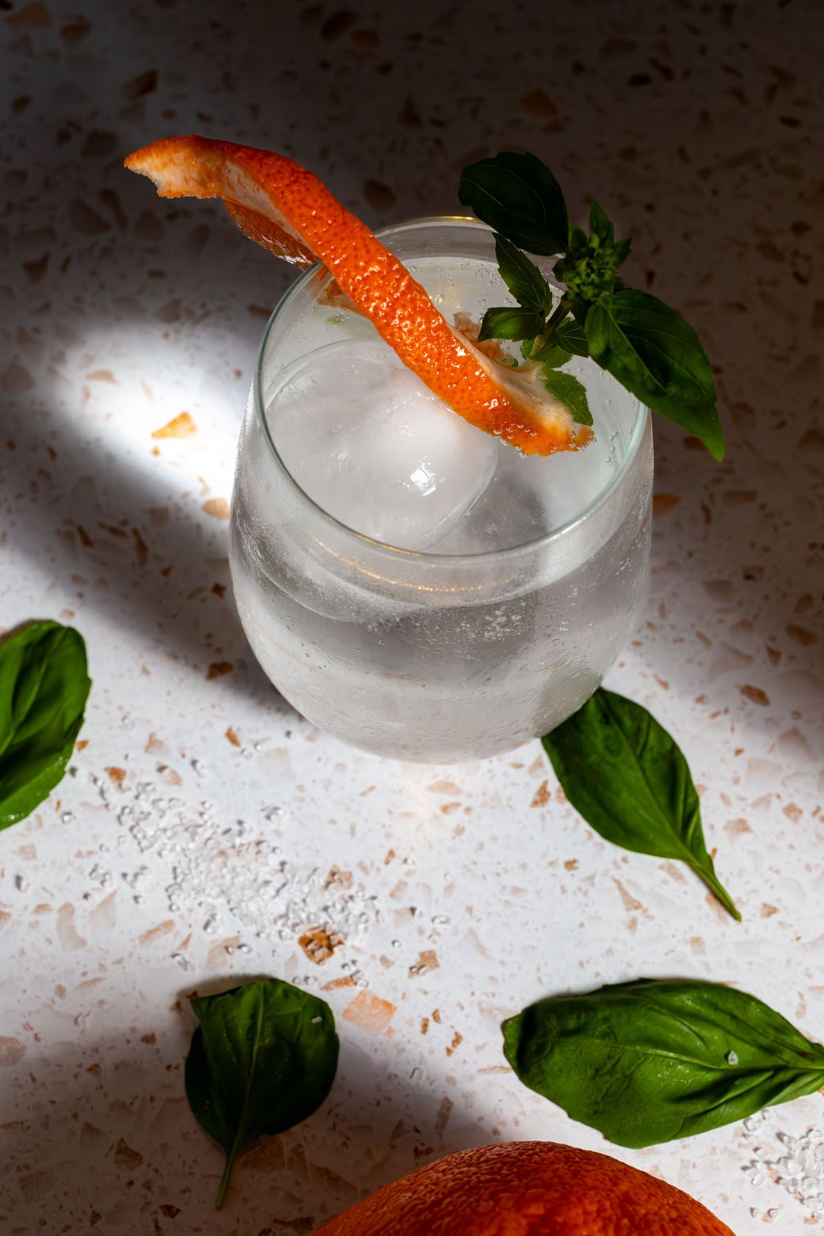 Fresh basil leaves and grapefruit citrus fruit with spritzer arranged on a white textured surface with dramatic lighting and shadows.