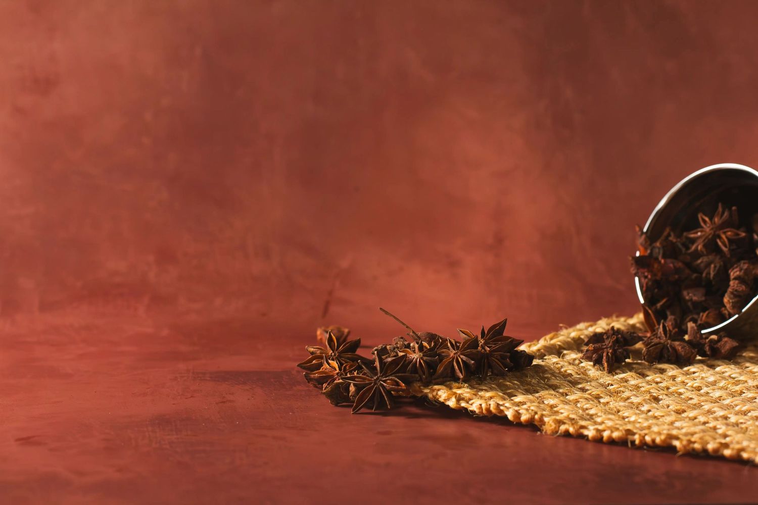 Scattered star anise spices spilled from a container onto a burlap fabric against a dark red background. Set design and food styling service