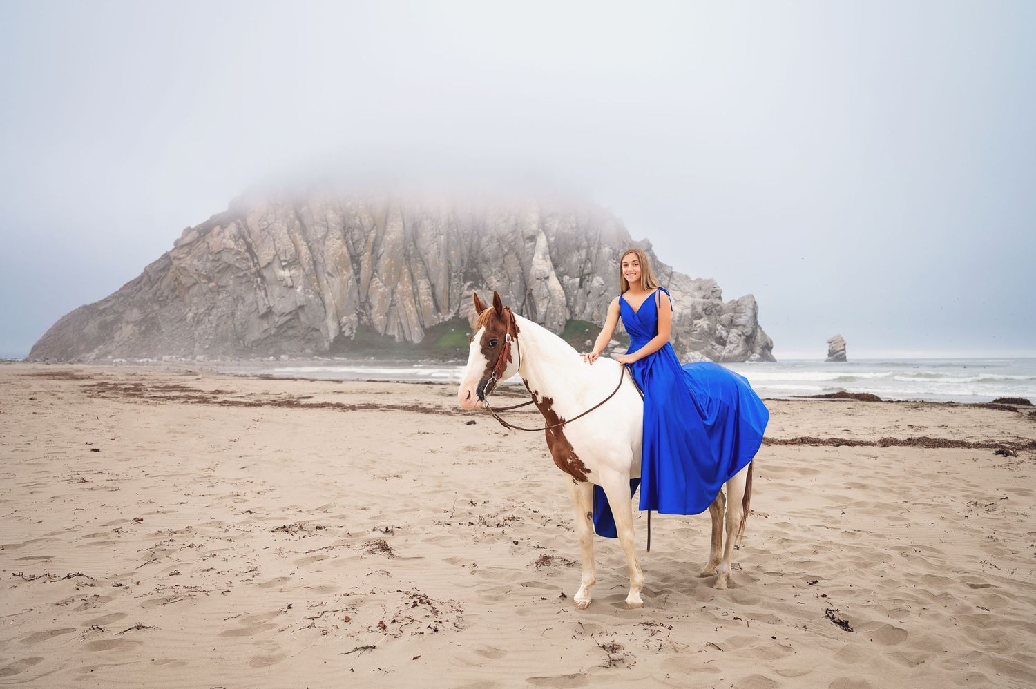 A figure in a royal blue dress rides a white horse along a foggy beach with majestic cliffs.