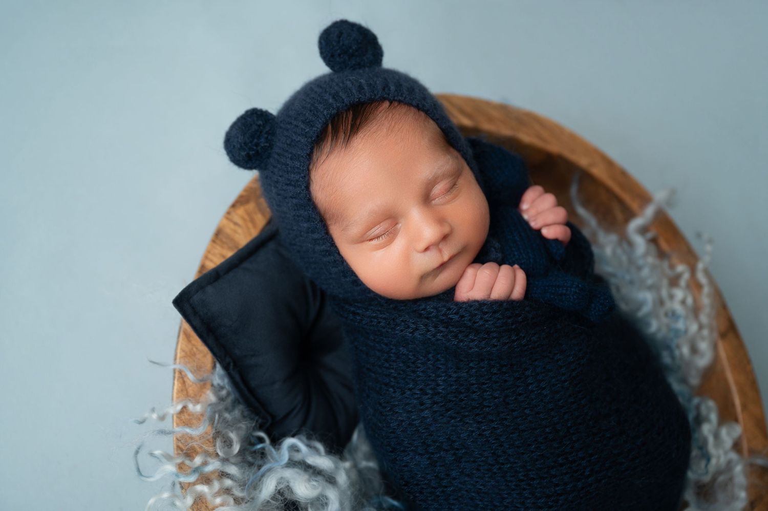 Sleeping newborn wrapped in navy knit blanket with bear ear hat rests in wooden bowl with gray fabric.