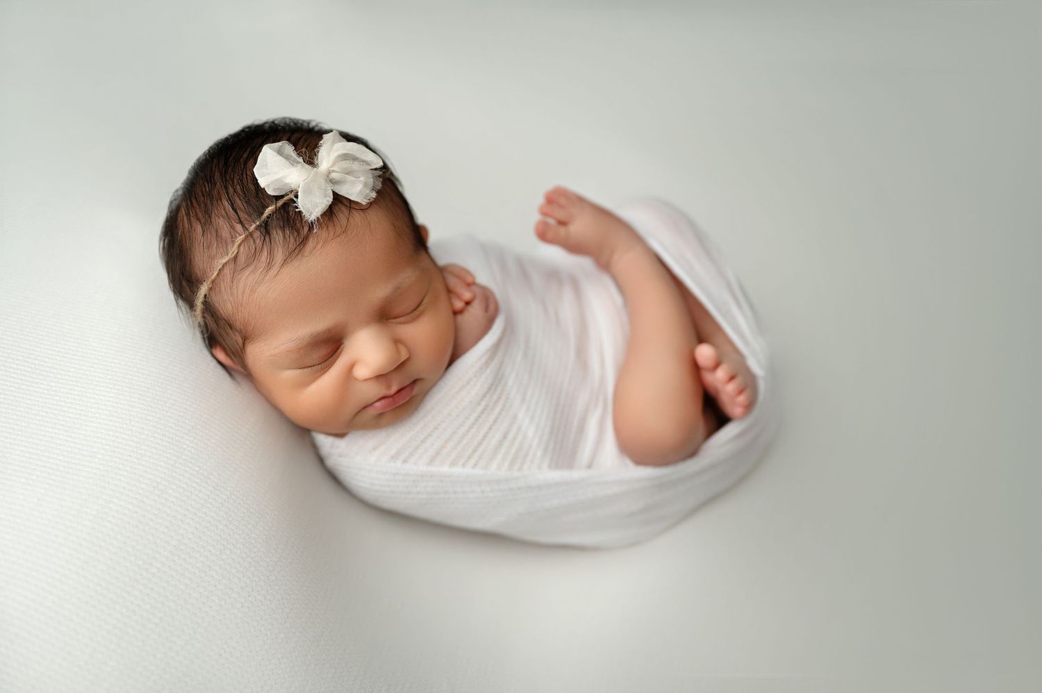 Sleeping newborn wrapped in white fabric with small bow headband.