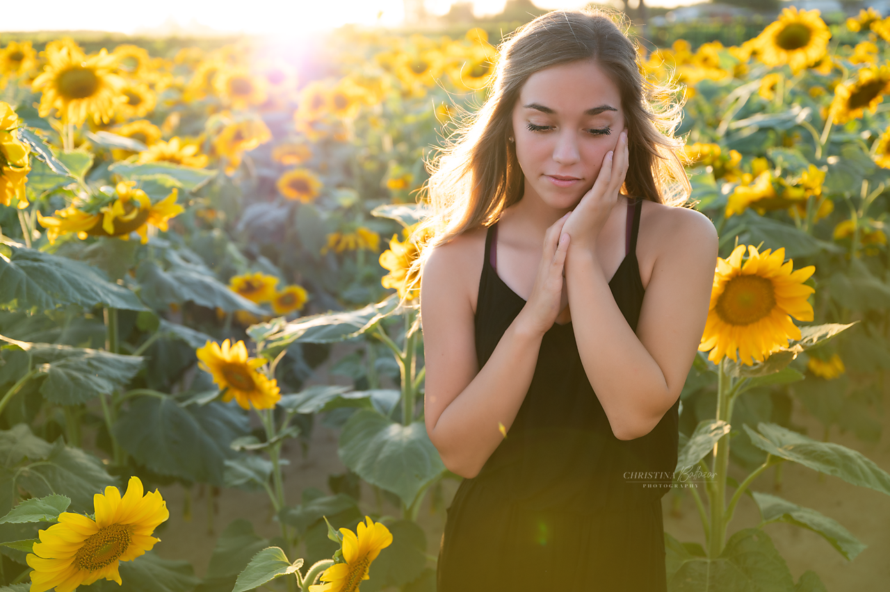 Person in black dress poses thoughtfully in sunflower field at golden hour.