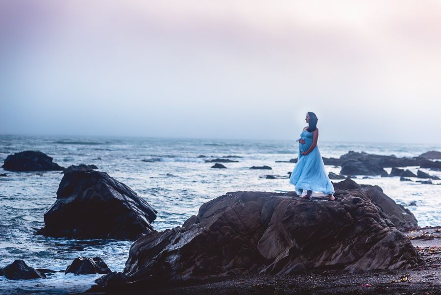 Silhouette of pregnant woman in blue gown standing on coastal rocks at dusk.