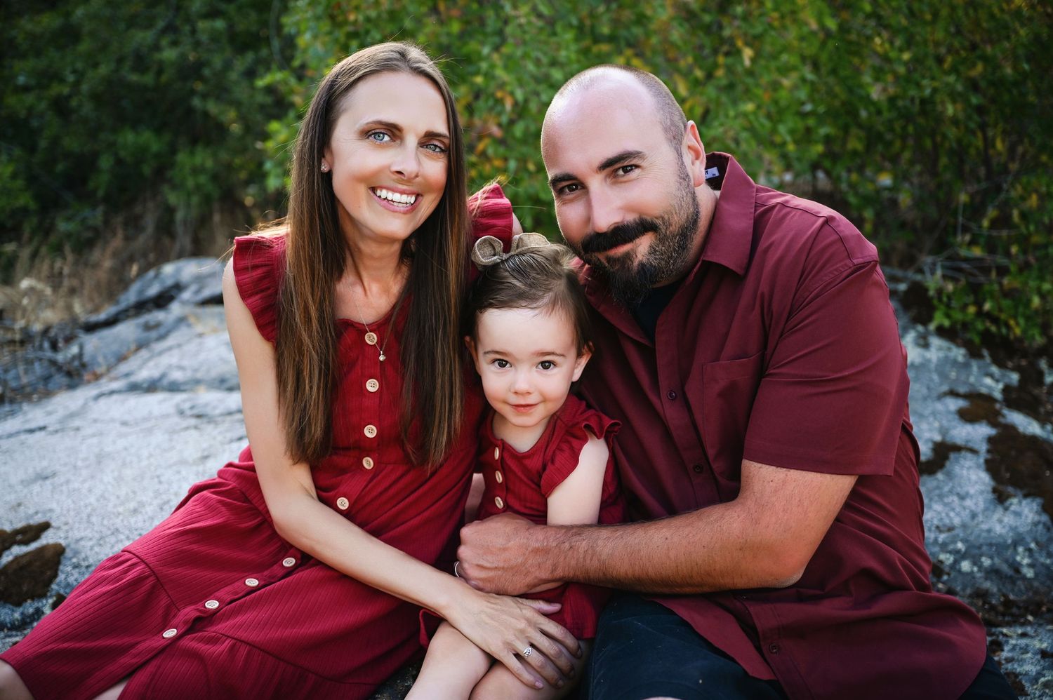 A family wearing burgundy colored outfits poses together outdoors for professional portraits in a natural setting. Fresno Family Photography