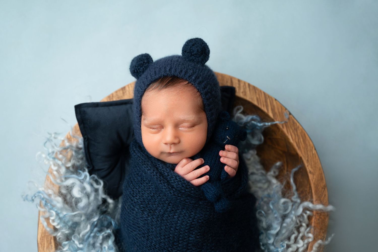 Baby wearing navy bear hat sleeps in wooden bowl with gray textured backdrop.