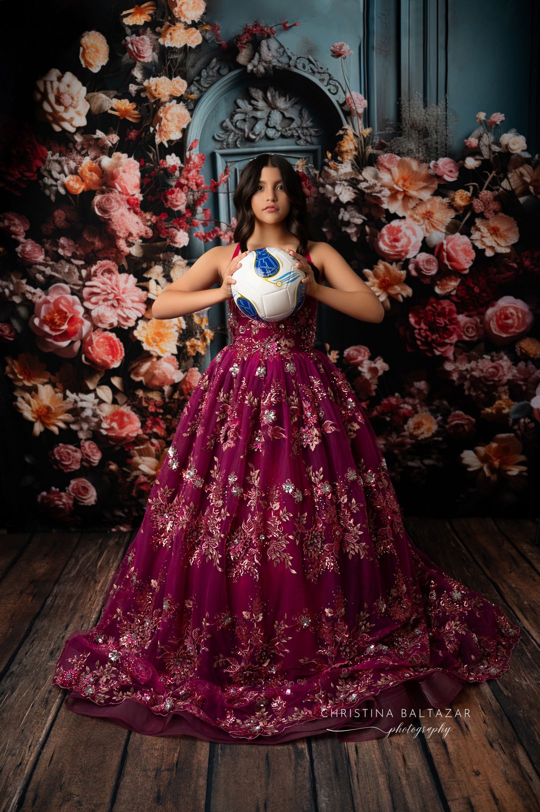 A whimsical portrait in a burgundy floral ballgown against a romantic backdrop of pink and peach roses.