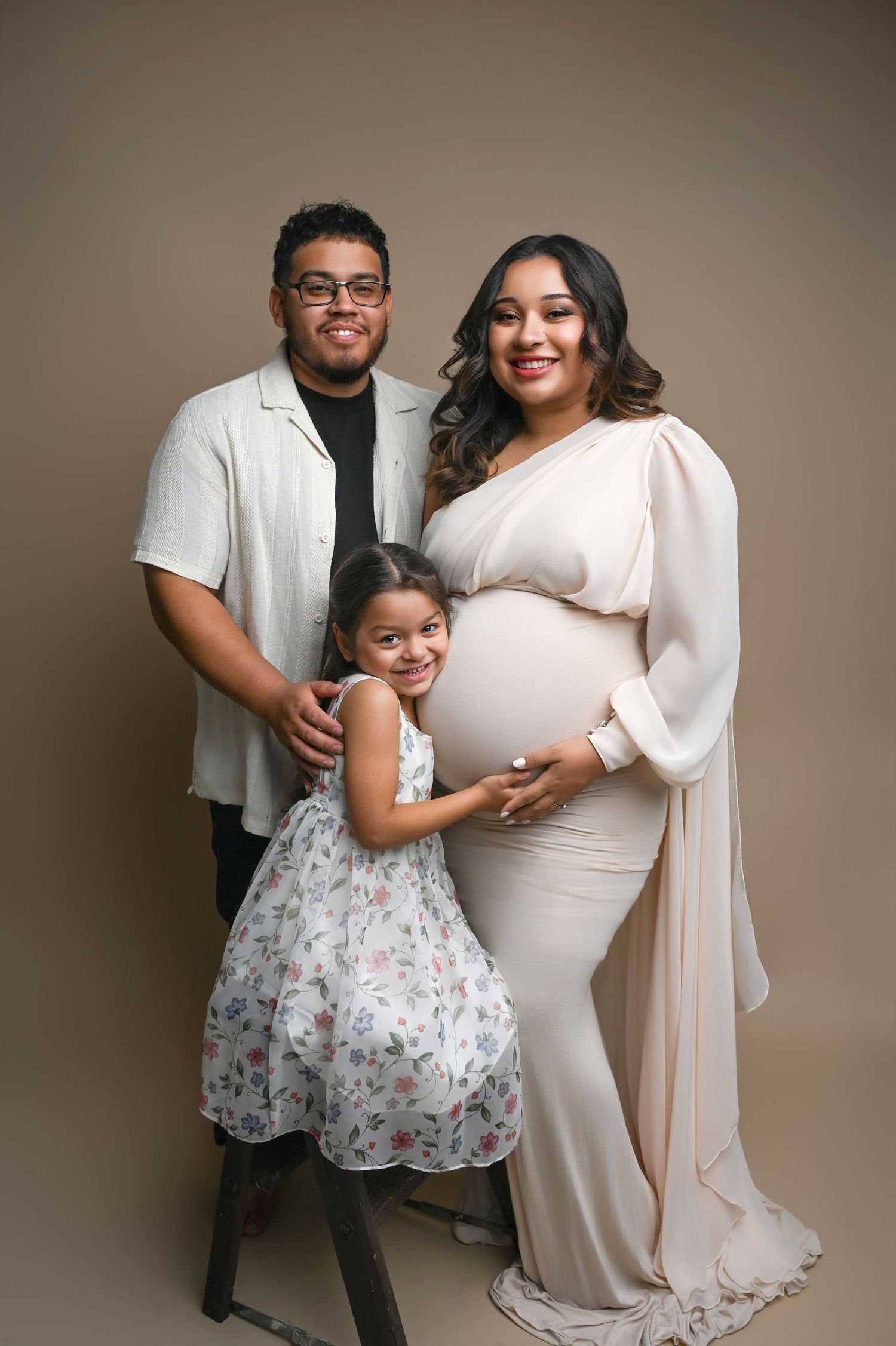 A family maternity photoshoot featuring a young girl in a floral dress and parents in white against a neutral backdrop.