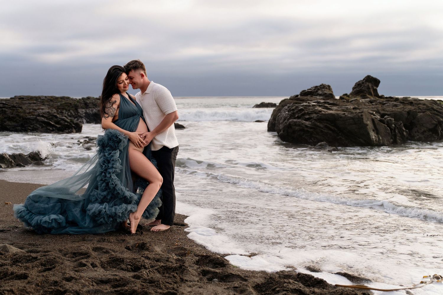 Couple embracing on rocky beach shore with dramatic cloudy sky and ocean waves.