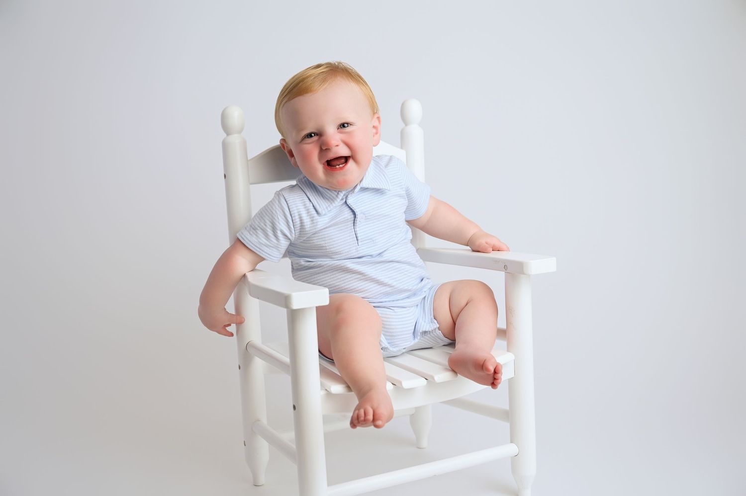 Baby in light blue outfit sits in white wooden chair during photo session.