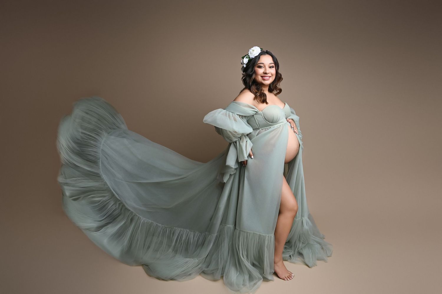 Pregnant woman in flowing sage green off-shoulder gown with floral crown poses gracefully against neutral backdrop.