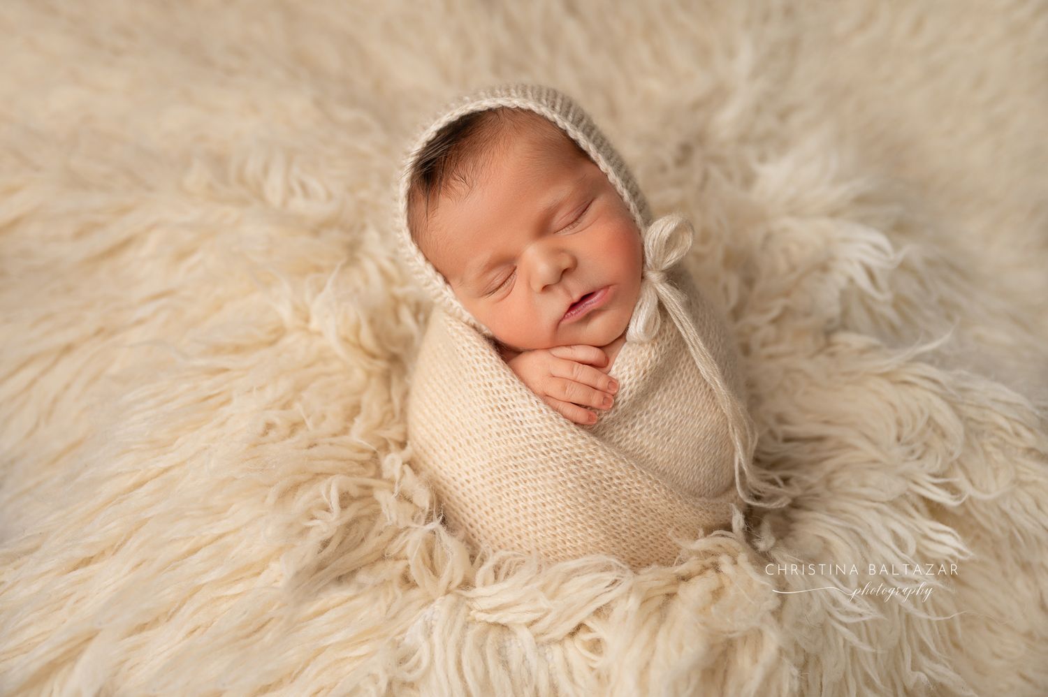 A peaceful newborn sleeping while swaddled in cream knit wrap on soft fluffy white fur backdrop. Fresno Newborn Photographer
