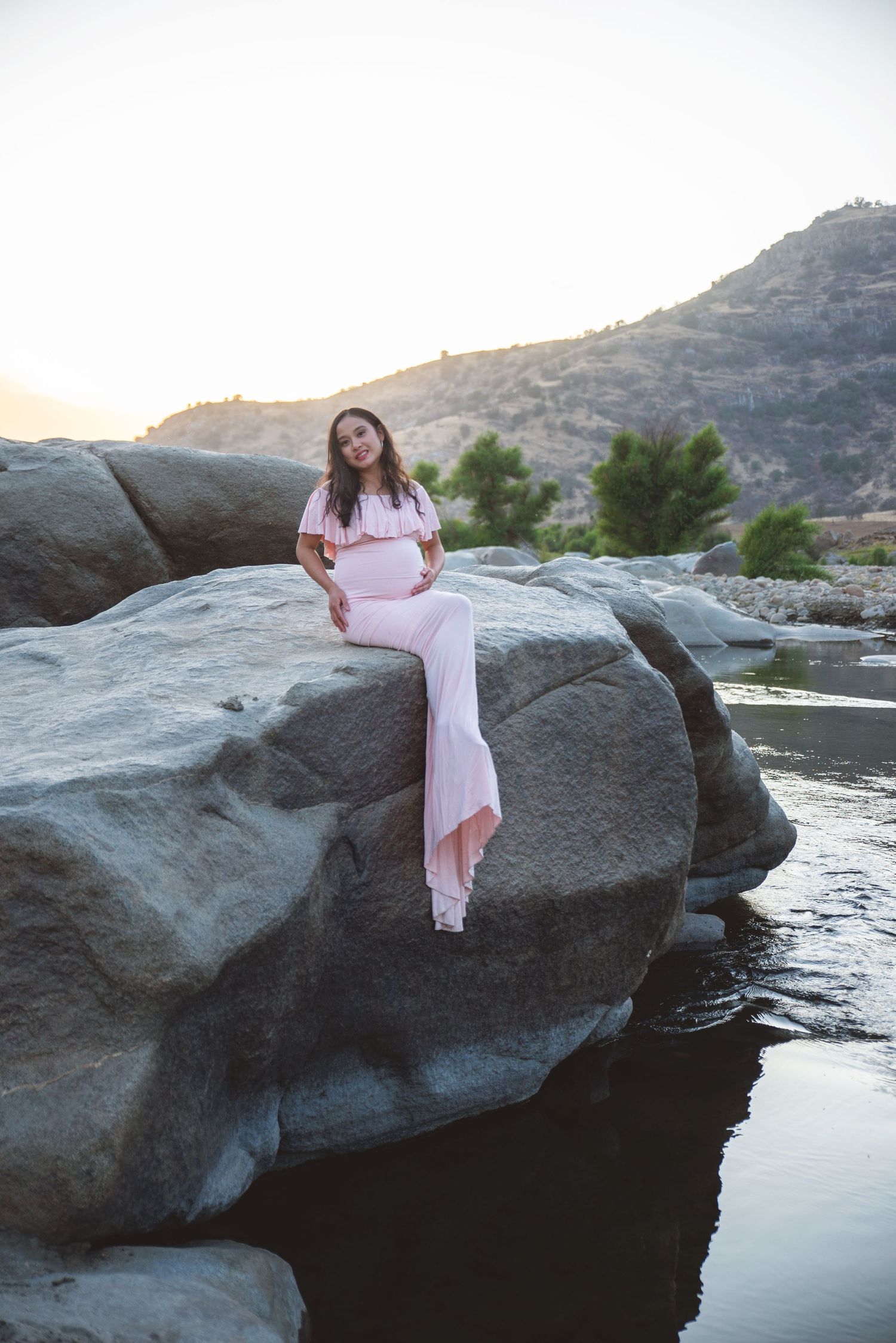 A maternity portrait in Three Rivers, CA of a woman in a pink dress sitting on a large rock near the river.