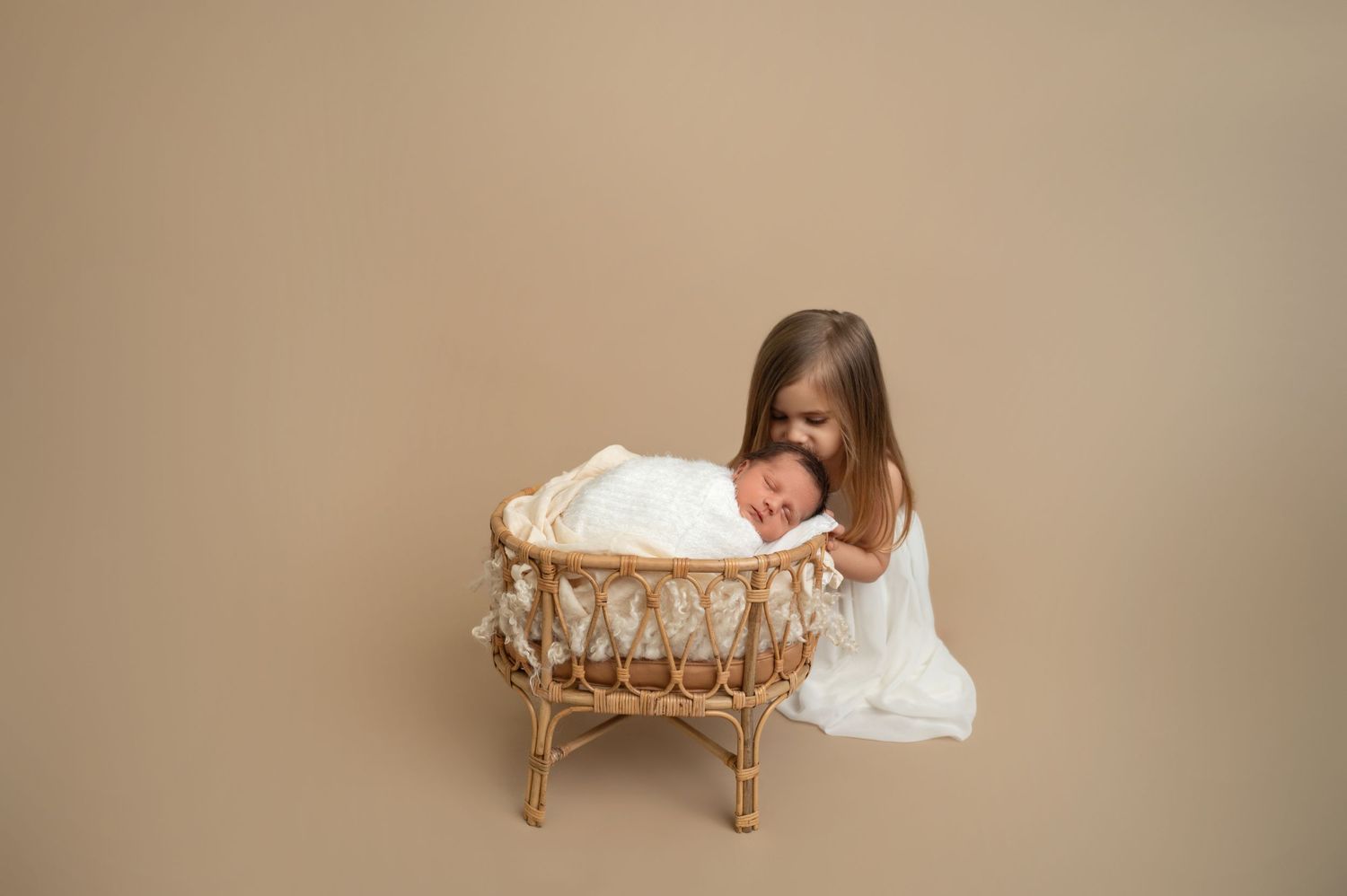 Child in white dress watching over newborn baby sleeping in woven bassinet.