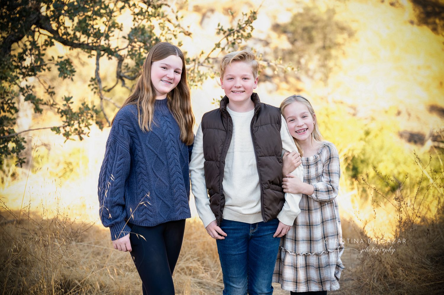 Group of three siblings stand together outdoors at golden hour in casual autumn attire. Hanford Family Photographer