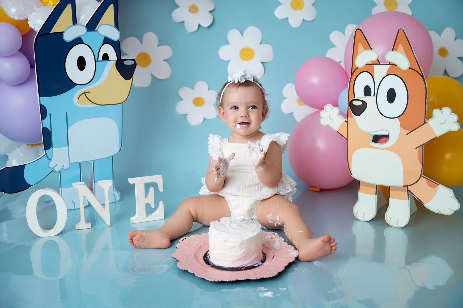 Whimsical first birthday setup with balloons, daisies and cartoon characters against blue backdrop.
