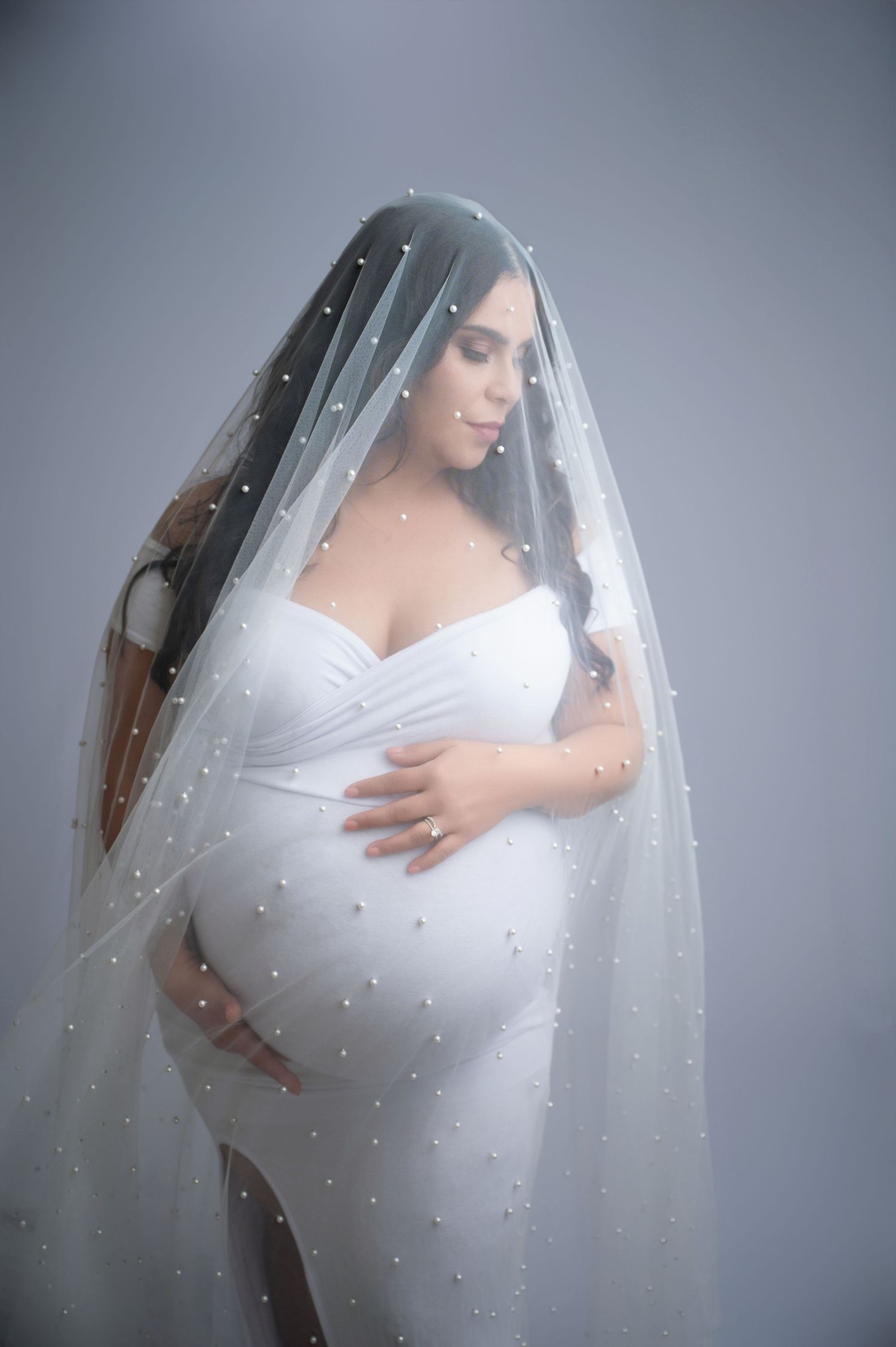 Ethereal maternity photos with flowing white dress and pearl-studded veil against grey background.