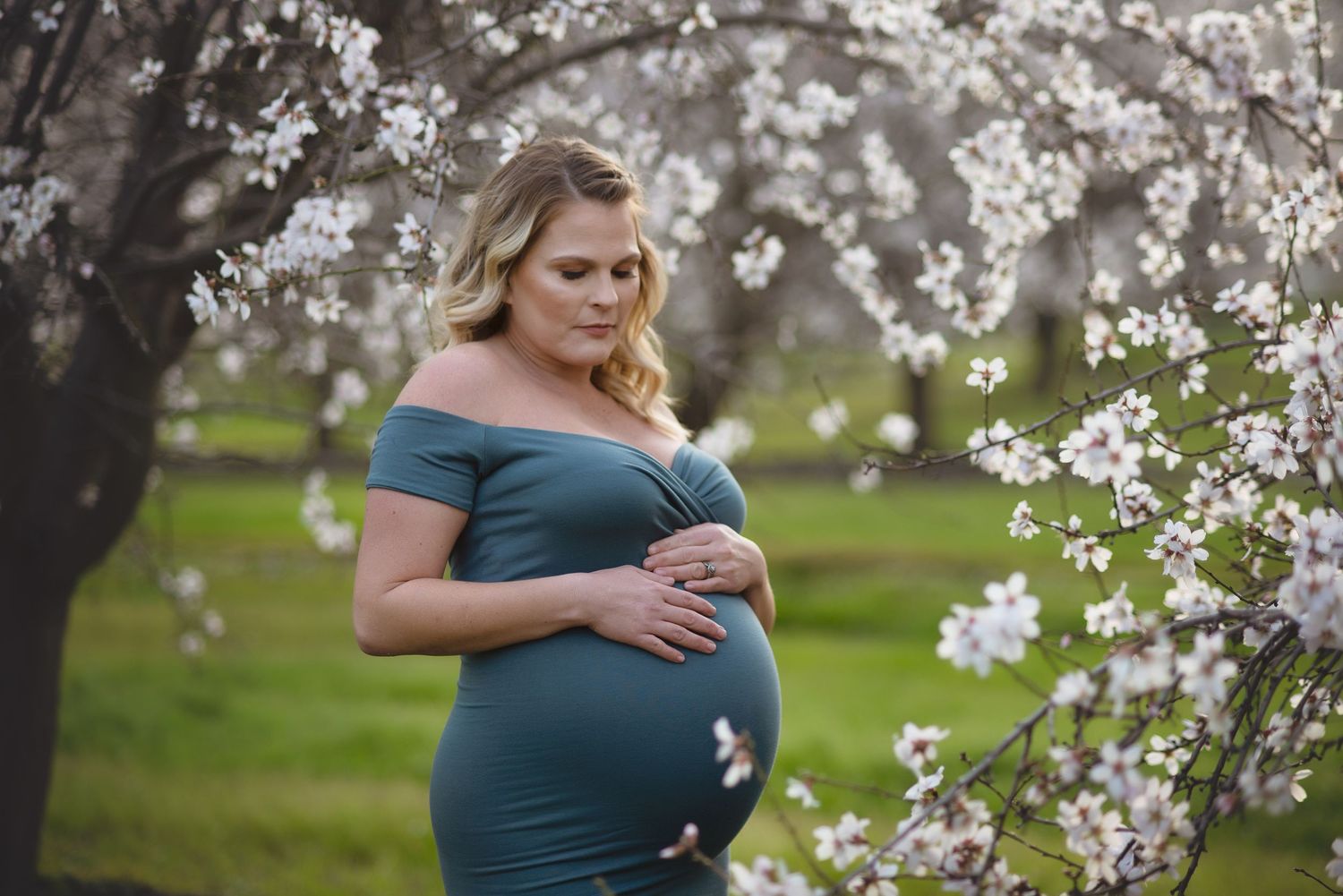 Expecting mother in a teal dress poses gracefully among blooming almond blossoms in Lemoore, CA for maternity portraits.