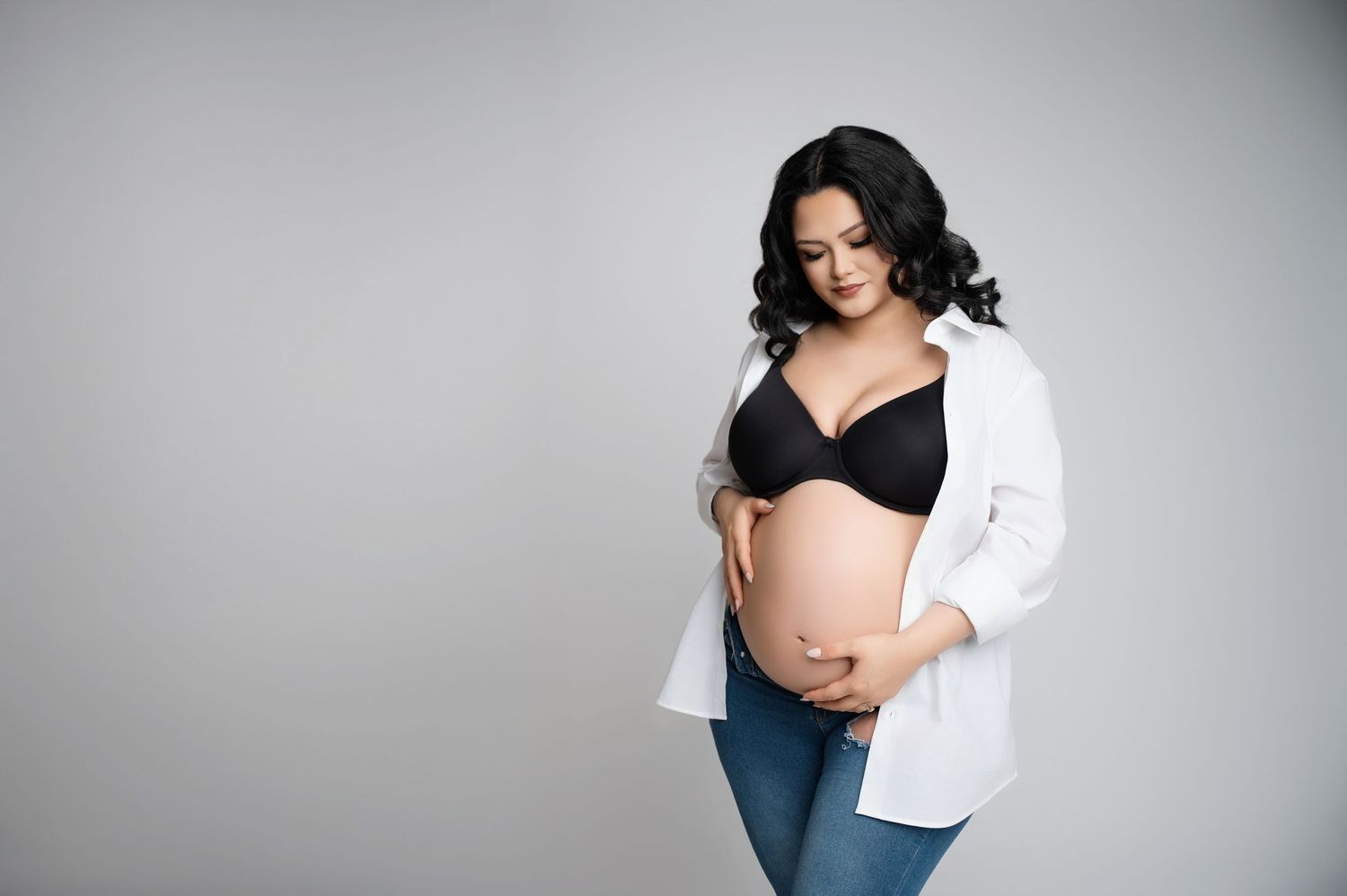 A pregnant woman wearing a black bra and open white shirt poses against a gray background for a maternity photo. Fresno Maternity Photos