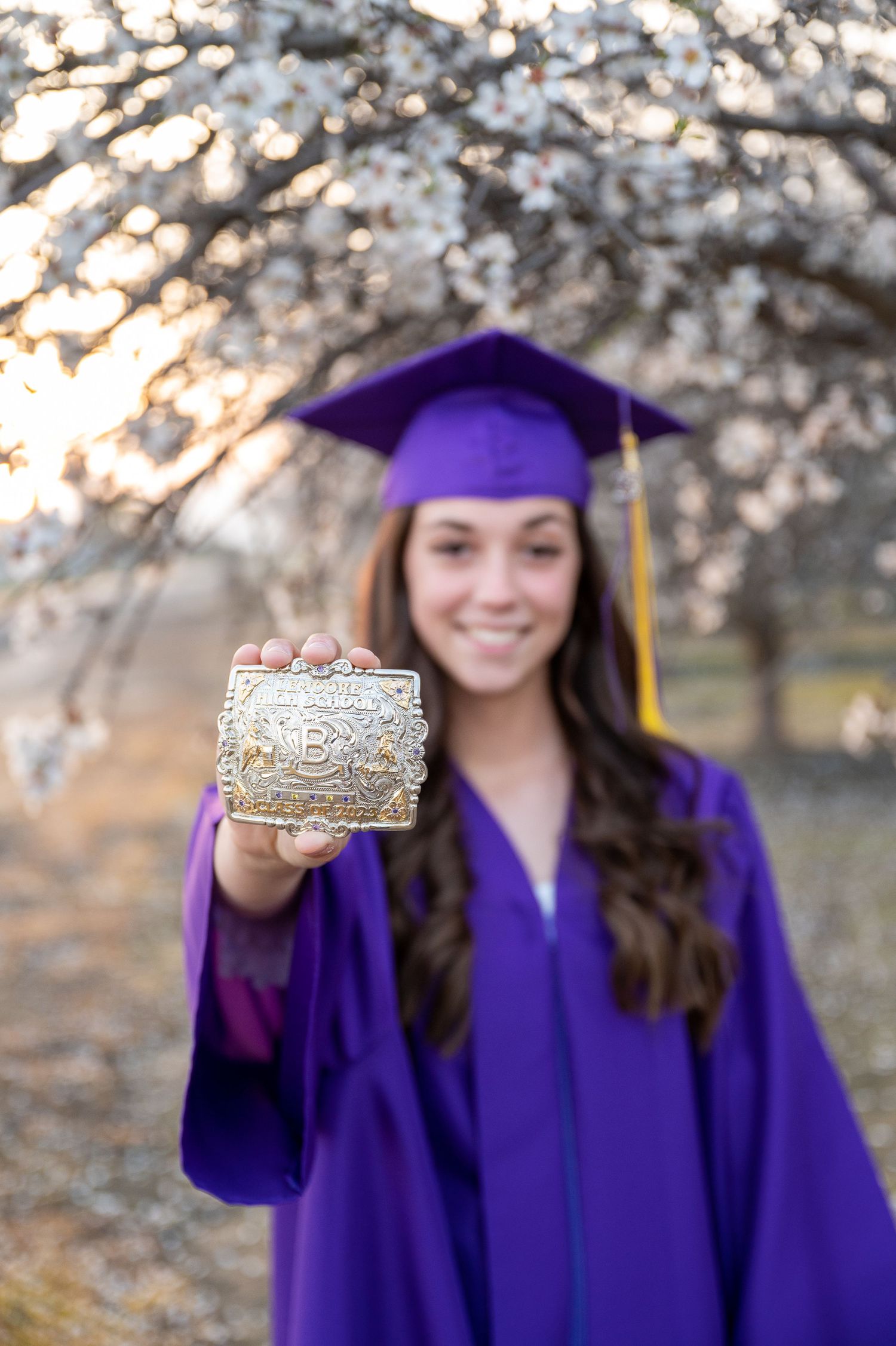 Graduate in purple regalia displays class ring among blooming spring trees.