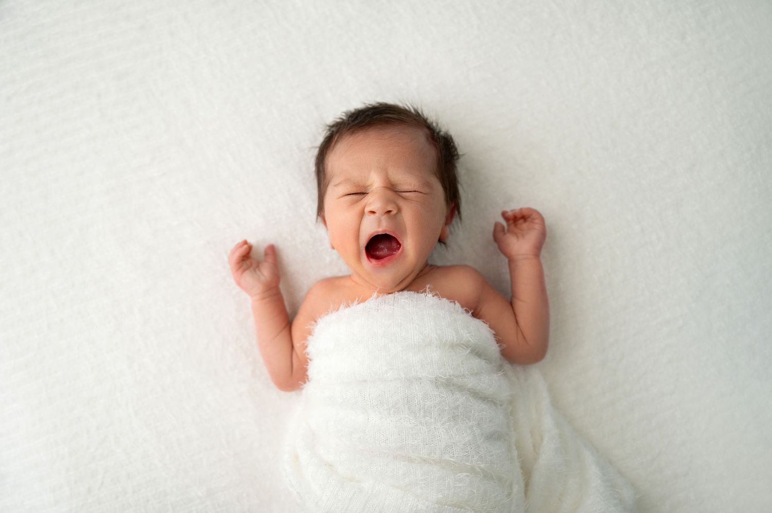 Newborn baby wrapped in white blanket yawning during professional photo session.