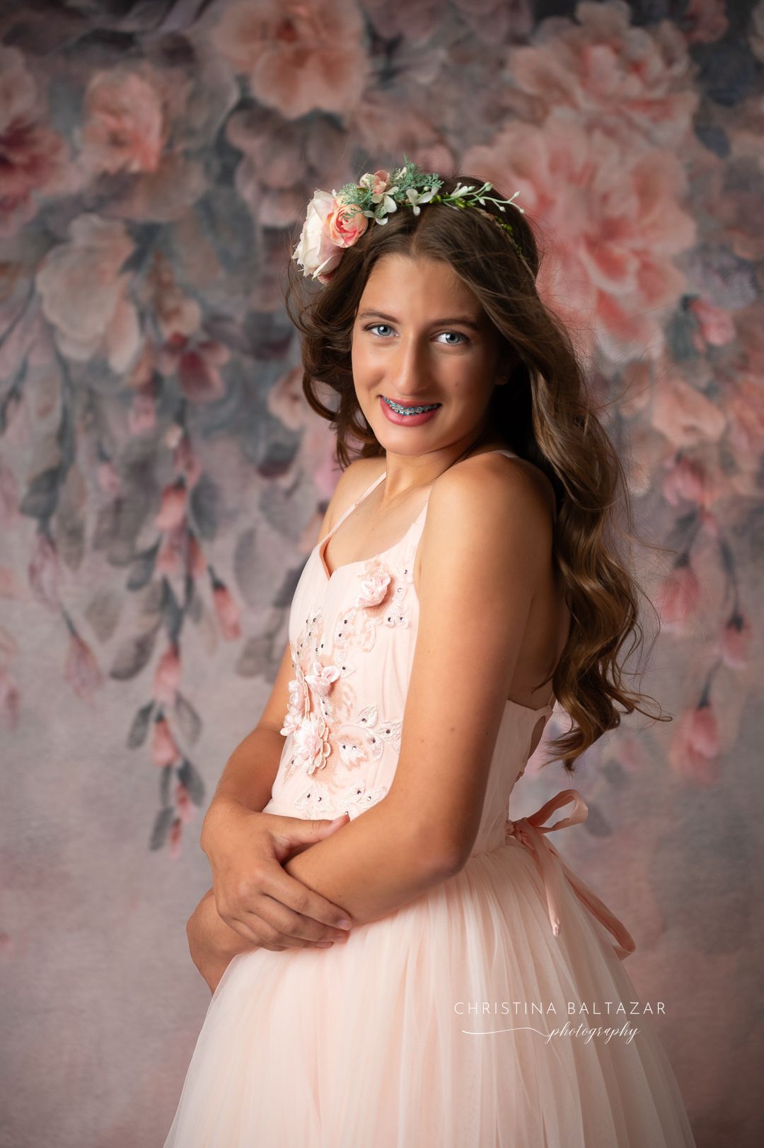 Portrait against floral backdrop featuring a long-haired person in pink tulle dress wearing delicate floral crown. Hanford Senior Photos