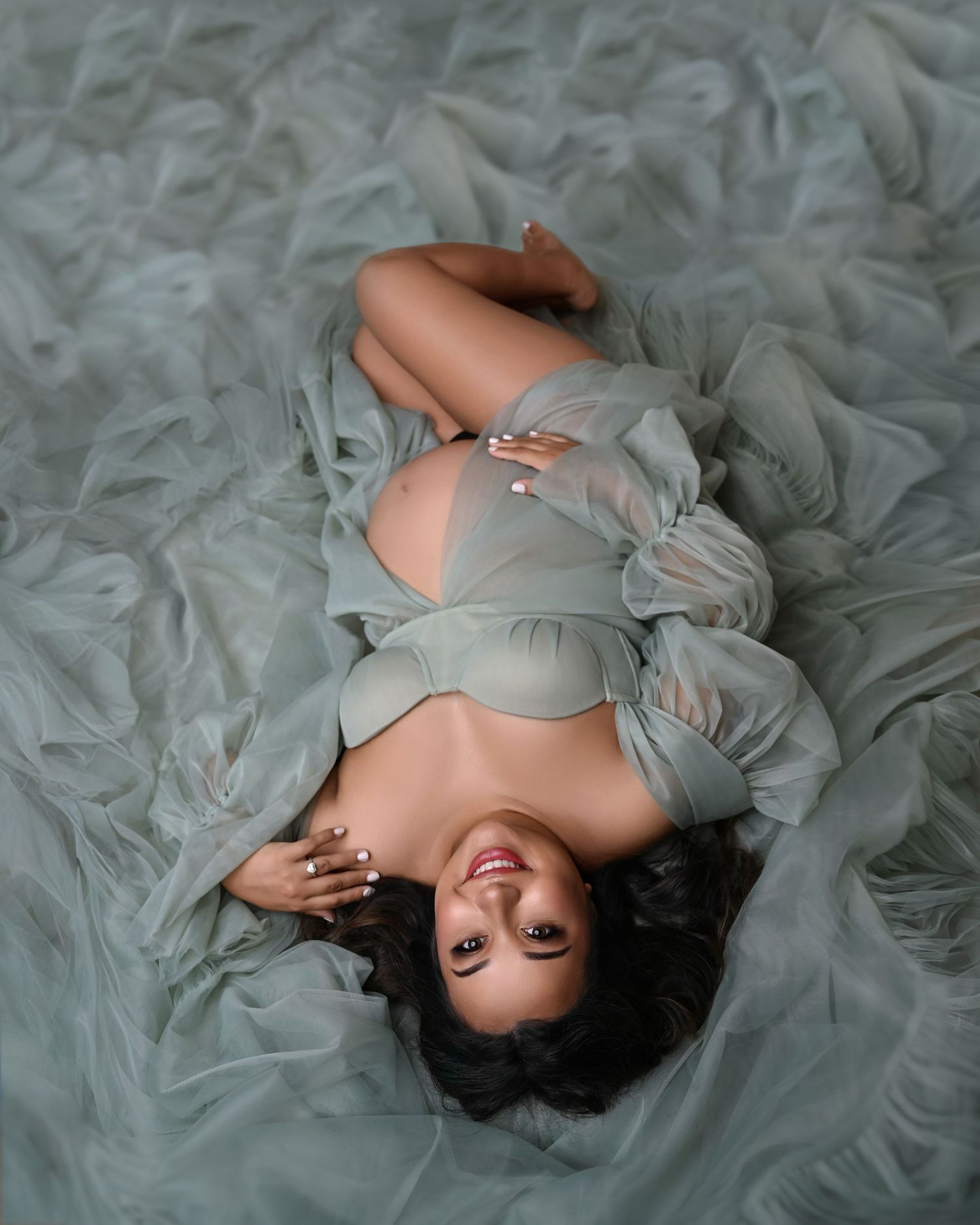 Woman in flowing gray dress lying gracefully on textured fabric in artistic maternity photoshoot. Hanford Maternity Photographer