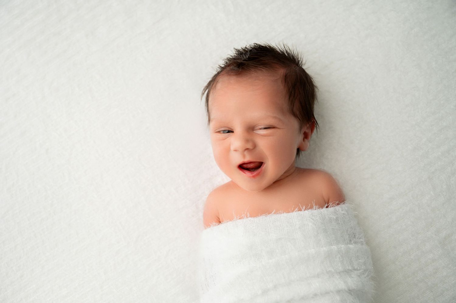 Smiling newborn wrapped in white fabric during professional portrait session.
