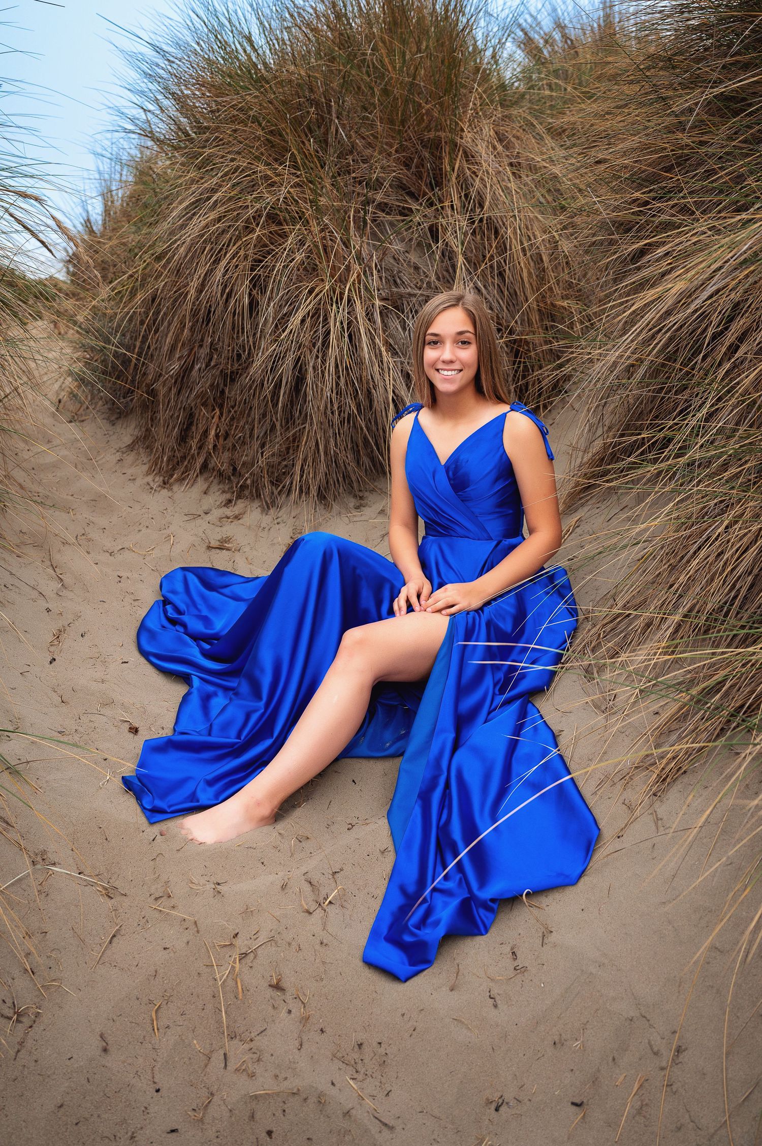 Flowing blue formal dress contrasts with beach dunes and coastal grass.