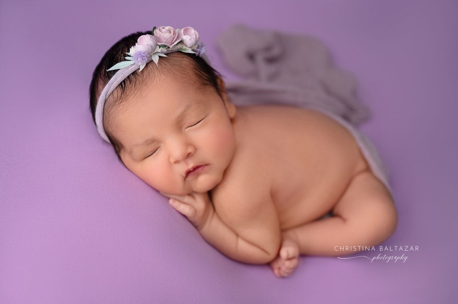 Sleeping newborn with white floral headband rests peacefully on purple backdrop during professional photo session. Fresno Newborn Photograph