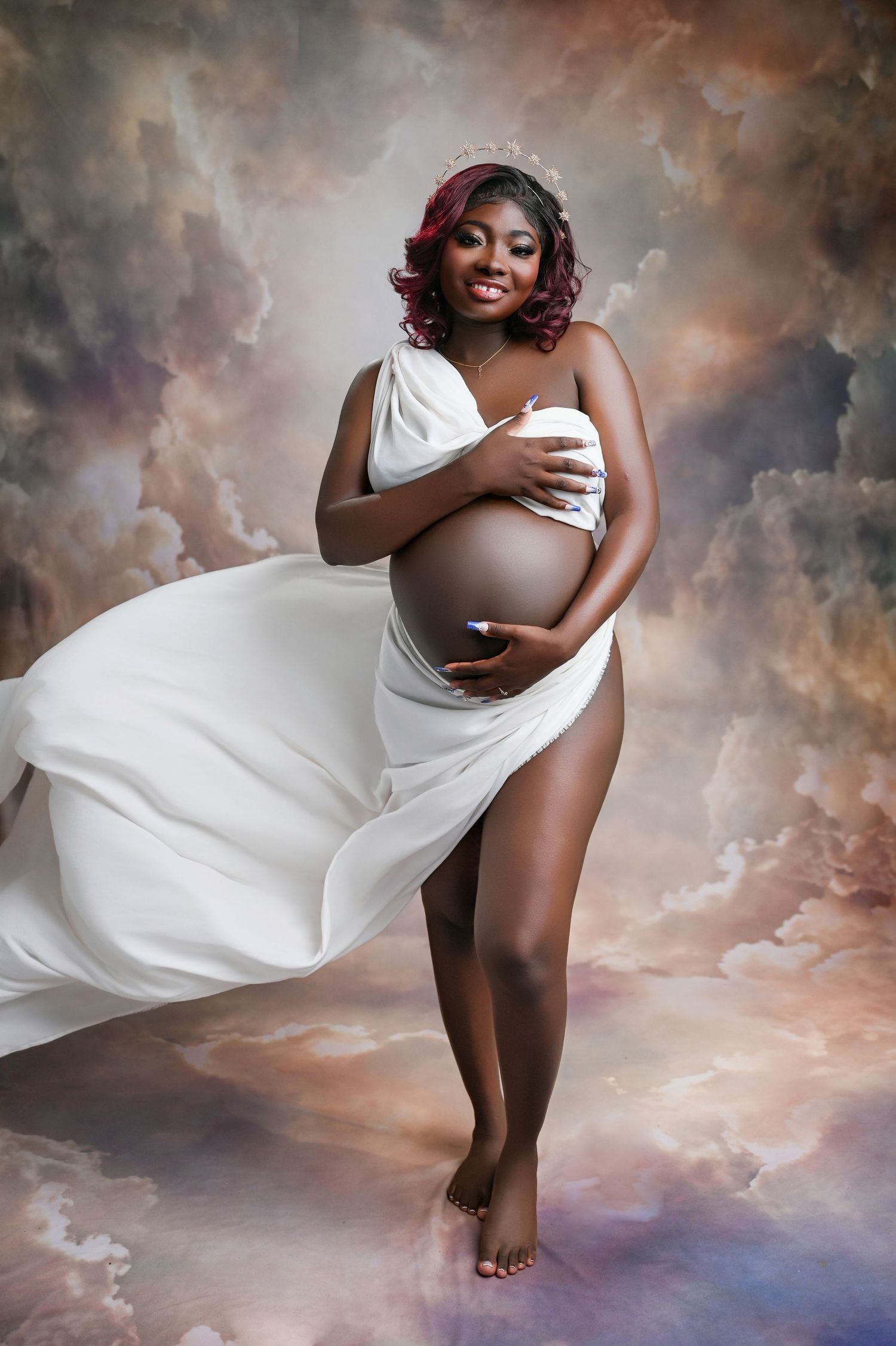 Graceful maternity photo with white dress flowing against ethereal cloudy background.