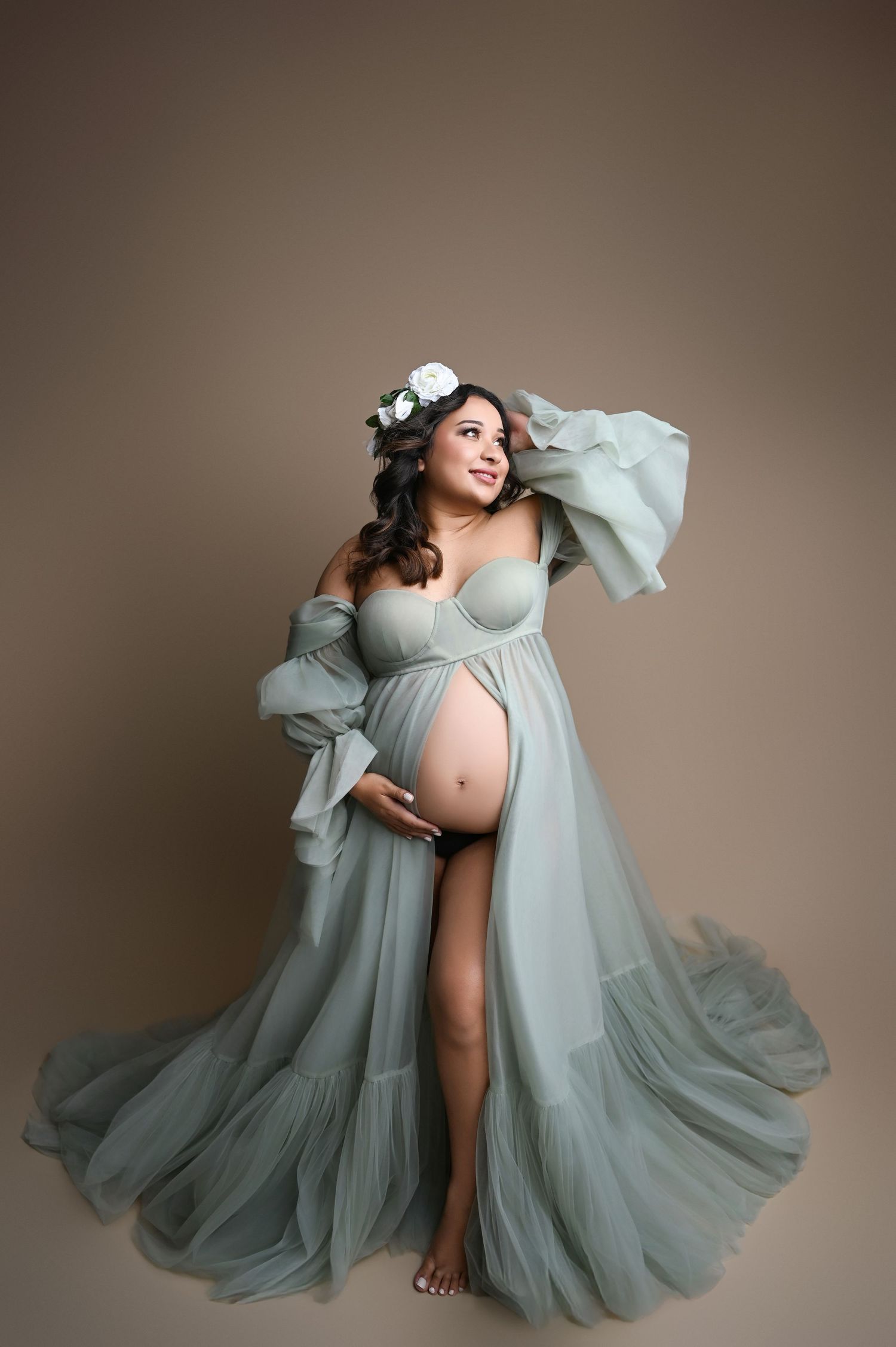 Artistic maternity portrait with flowing sage green tulle dress against brown backdrop.