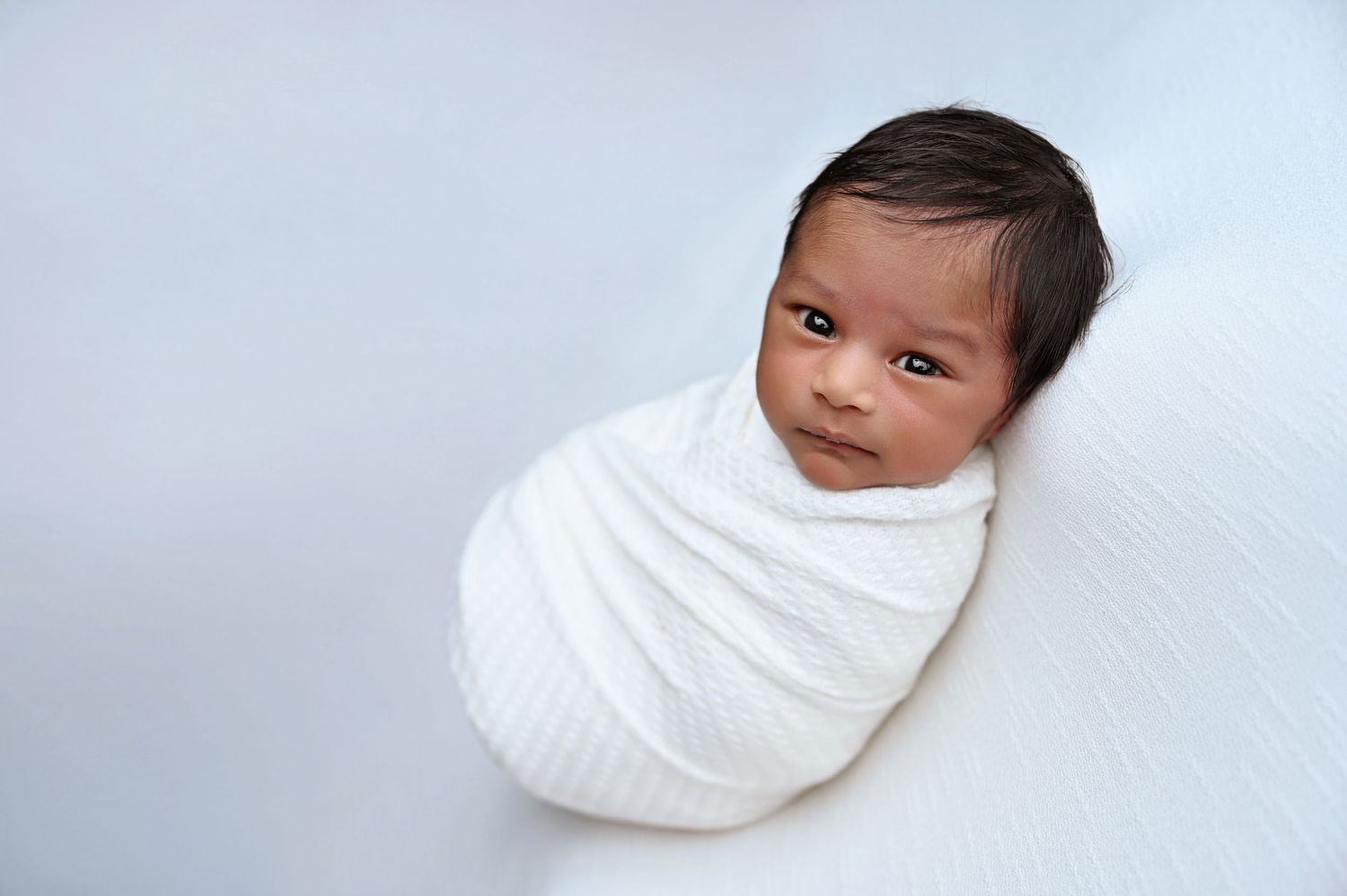 Newborn baby wrapped in soft white swaddle against light background.