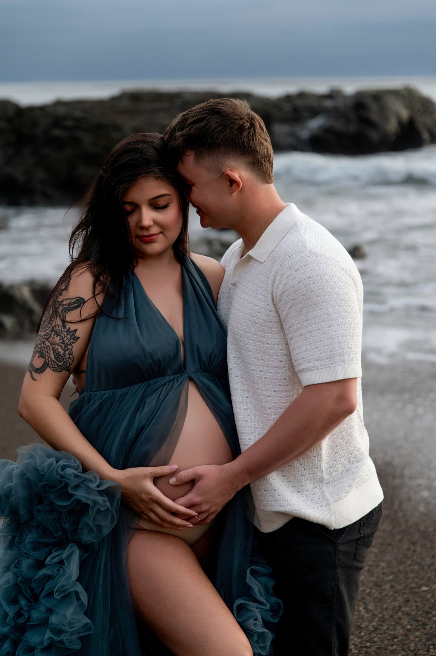 Intimate maternity photos captured on moody beach with flowing blue dress and white shirt.
