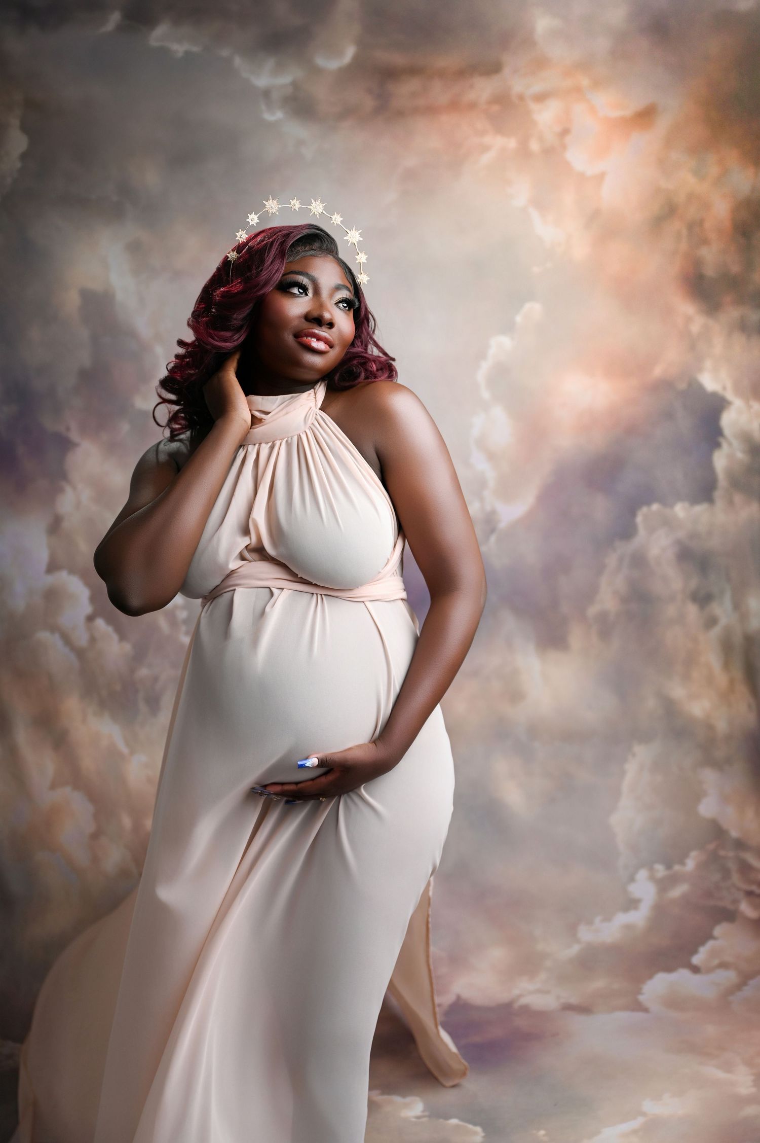 Ethereal maternity portrait against dreamy clouded backdrop wearing flowing white gown.
