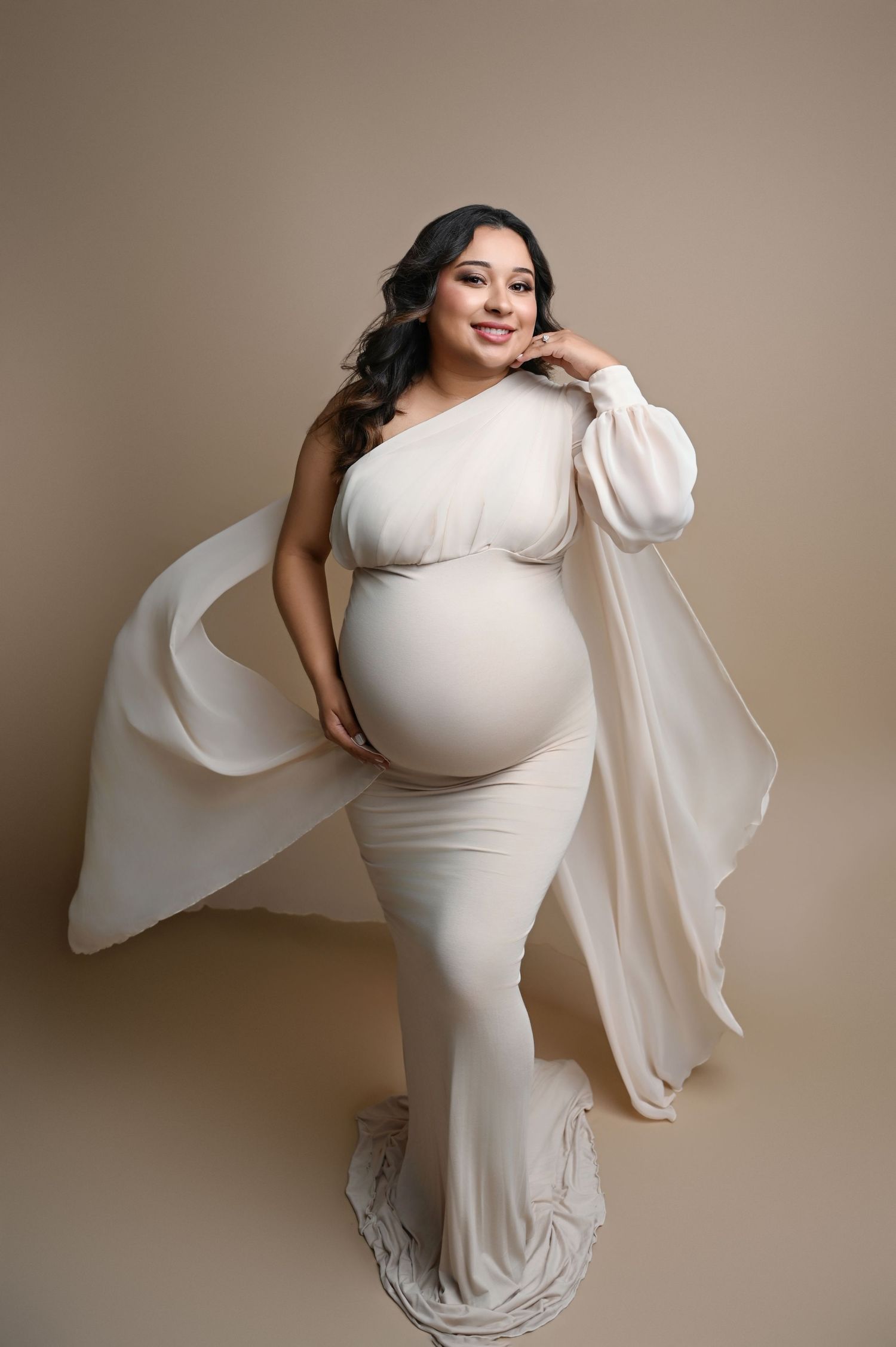 Elegant maternity photos in cream dress with flowing sleeves against beige background.