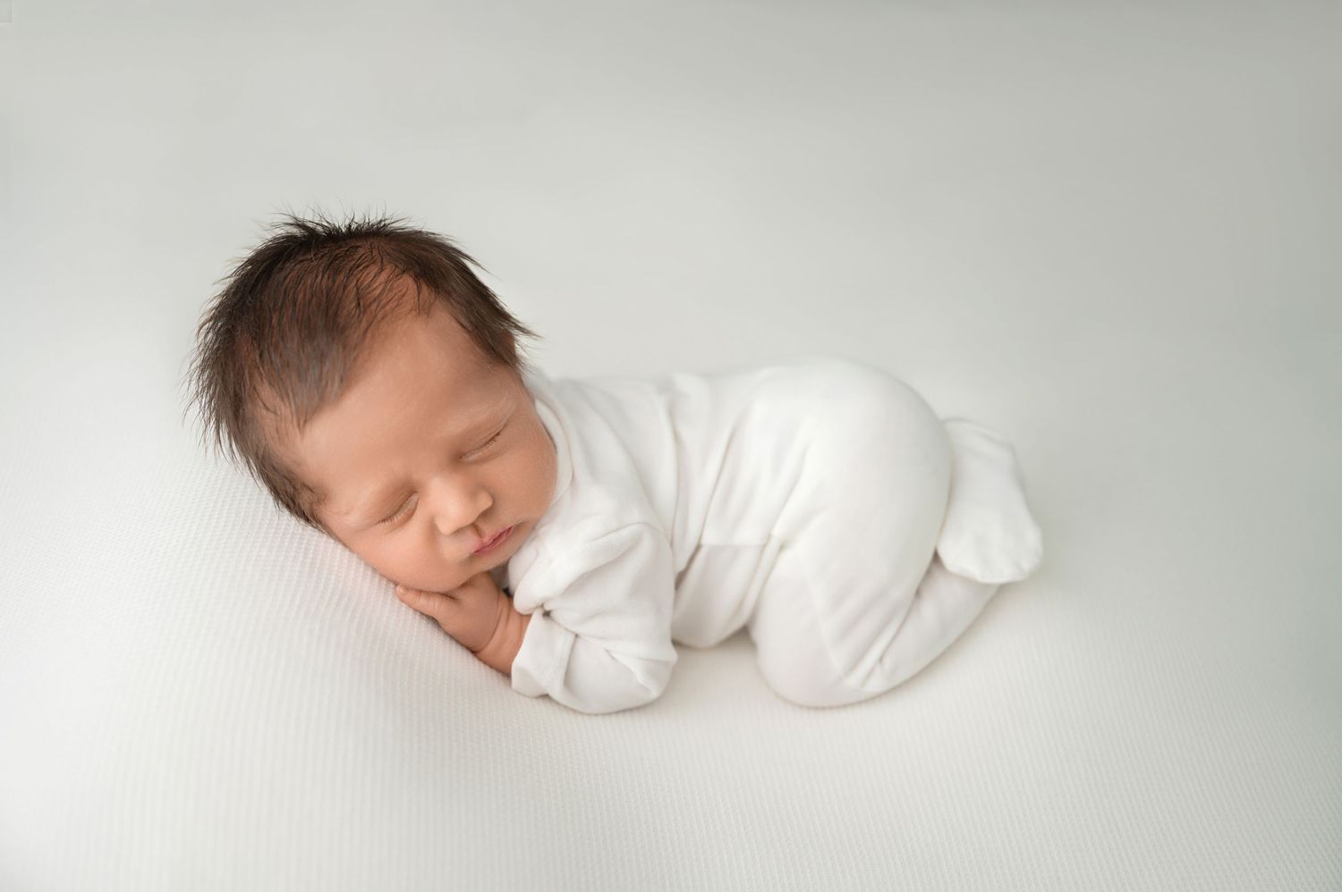 Sleeping newborn in white outfit curled up on soft white background in studio.