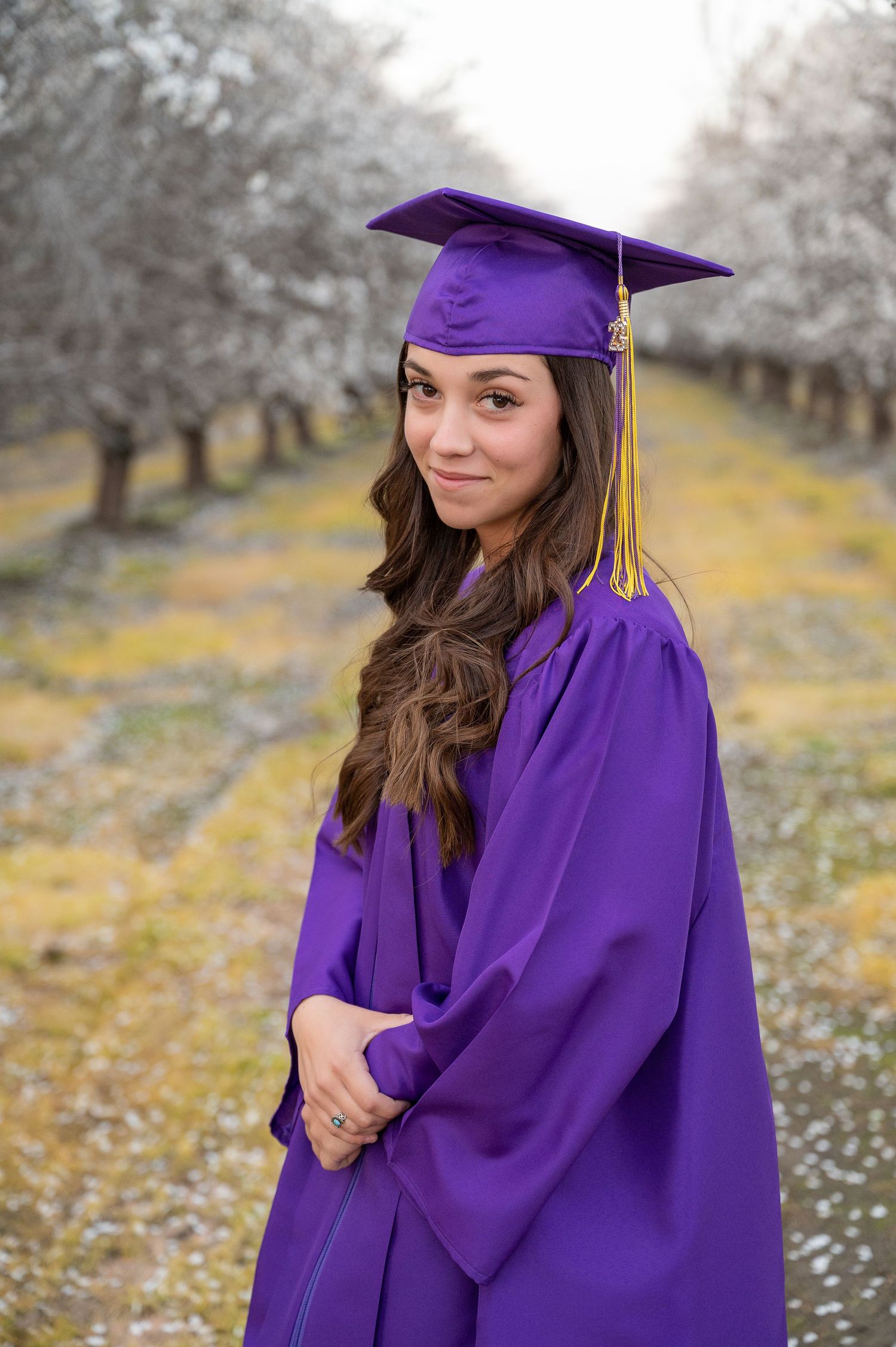 Purple graduation regalia stands out against white spring blossoms in an orchard setting.