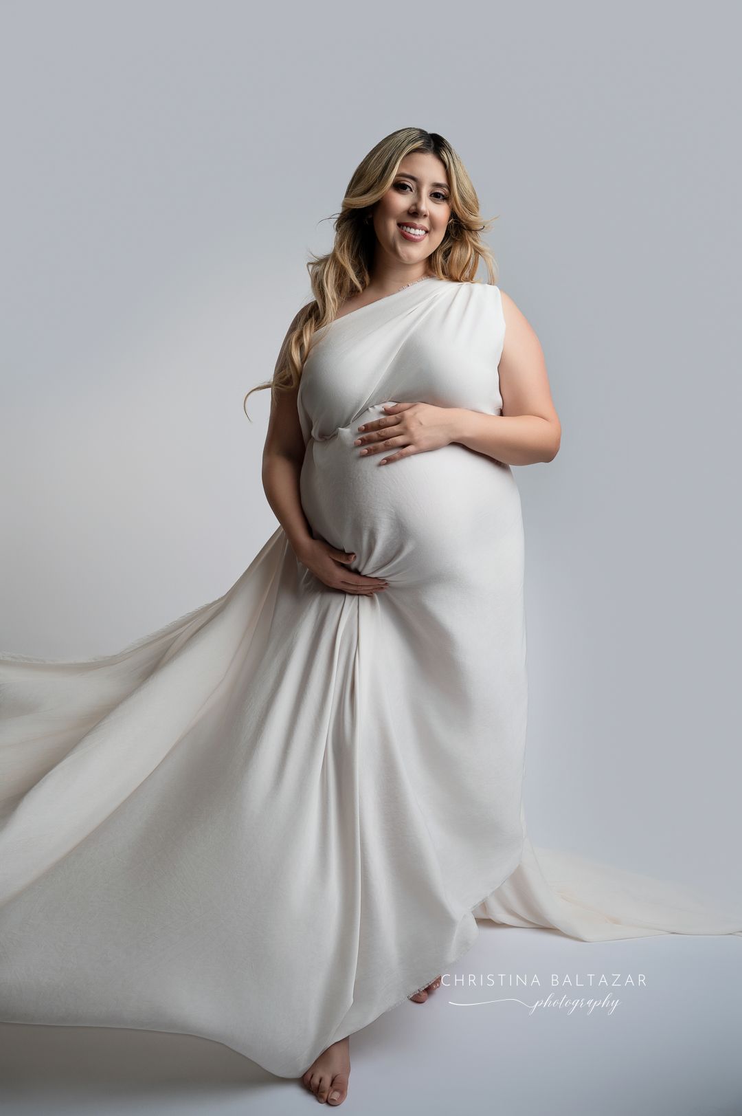 A pregnant woman in a flowing white maternity dress poses gracefully against a light gray backdrop for a studio portrait. Fresno Maternity