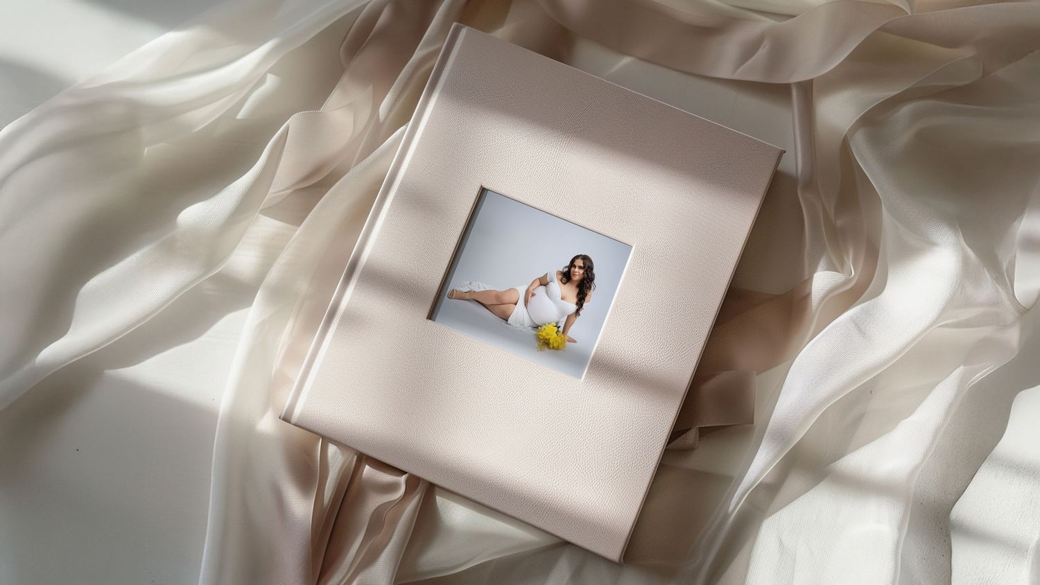 An elegant maternity photo album with cream leather cover placed on silky white fabric in soft natural lighting. Fresno Maternity Photos