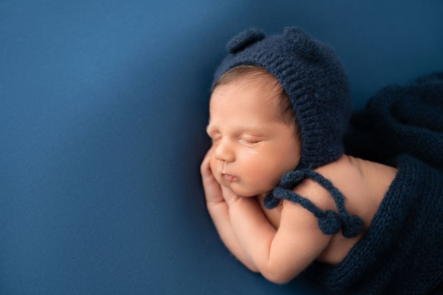 Newborn wearing knit bonnet and wrap sleeps peacefully against blue backdrop.